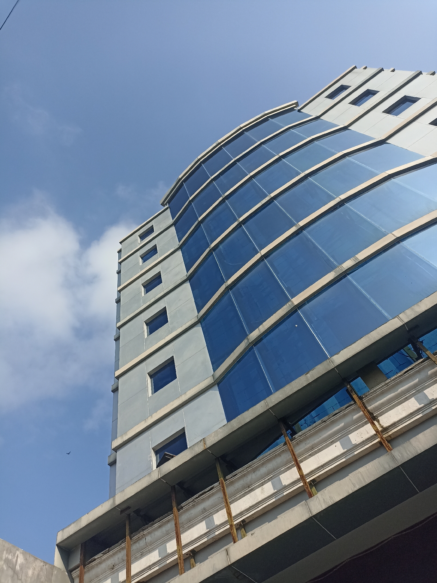 The image shows a low-angle view of a modern, curved building with a facade of blue glass and grey panels against a bright blue sky with scattered white clouds. The building's design features horizontal bands of glass interspersed with smaller, rectangular windows on the grey sections. In the foreground, a section of an older, unfinished or partially constructed structure is visible, with exposed concrete and rusty support beams. A single bird is in flight in the upper left portion of the sky. The scene appears to be an urban environment, likely in Mojopanggung, Indonesia, given the context provided.