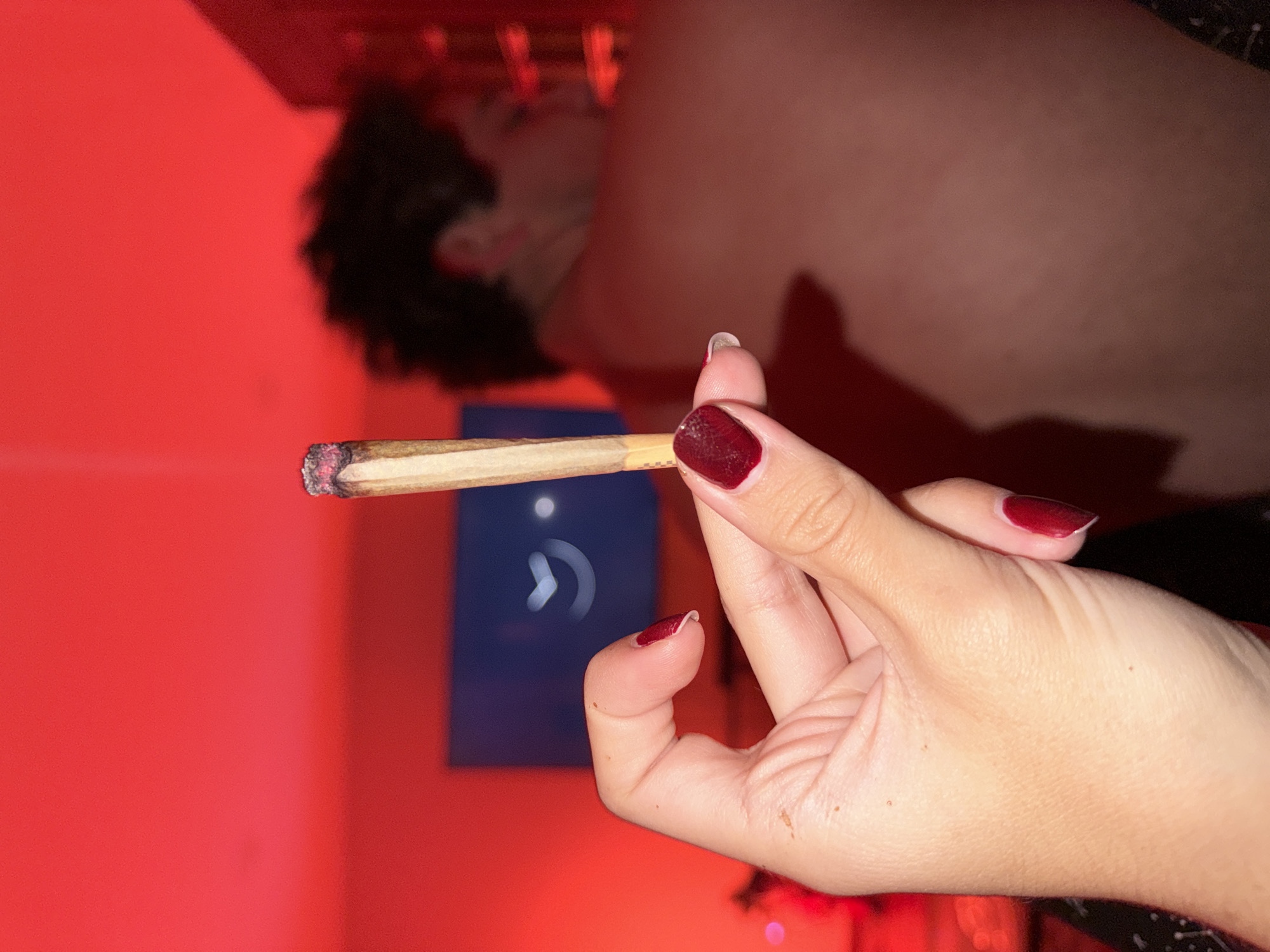 A hand with dark red nail polish holds a lit marijuana cigarette in front of a red wall. A person's blurred face and torso appear in the background. A blue object with a white curved arrow is in the background. The location is unknown.