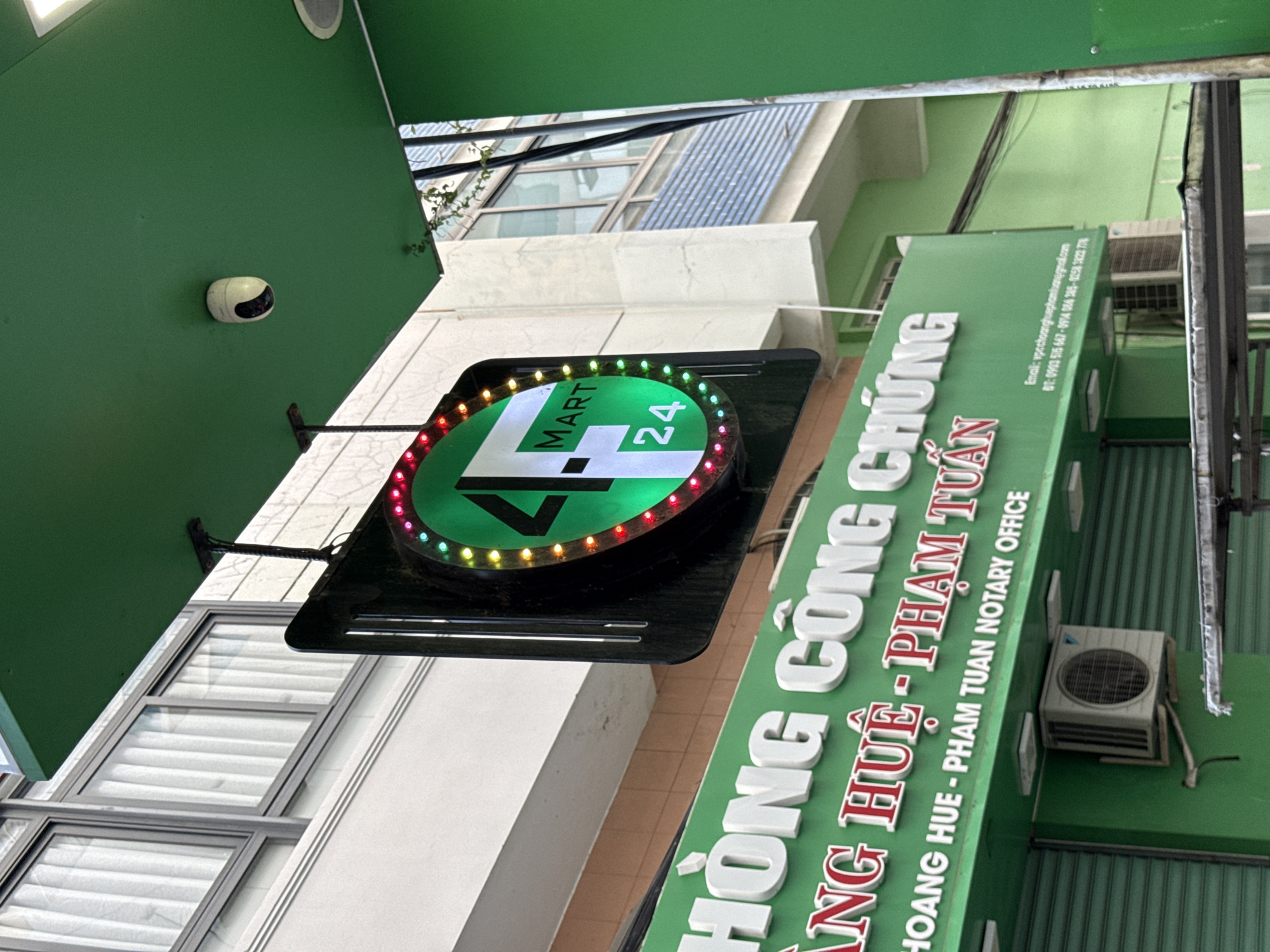The image shows the exterior of a business in Nha Trang, Vietnam. A prominent green sign with white, raised lettering reads "HỒNG CÔNG CHỨNG", and below it "ANG HUỆ - PHẠM TUẤN NOTARY OFFICE". The sign also contains smaller text detailing an email address and phone numbers.
Above the green sign, a round, black-bordered sign is suspended, featuring a green and white logo with the text "4 MART 24". This sign is illuminated by a ring of colorful LEDs. A small, white security camera is visible mounted on a green overhang above and to the right of the "4 MART 24" sign.
To the left of the signs, a building with large windows and white curtains is visible. The lower part of the image shows green roll-up shutters, characteristic of storefronts. An air conditioning unit is mounted on the green wall below the notary office sign. The overall scene suggests a commercial area in the city.