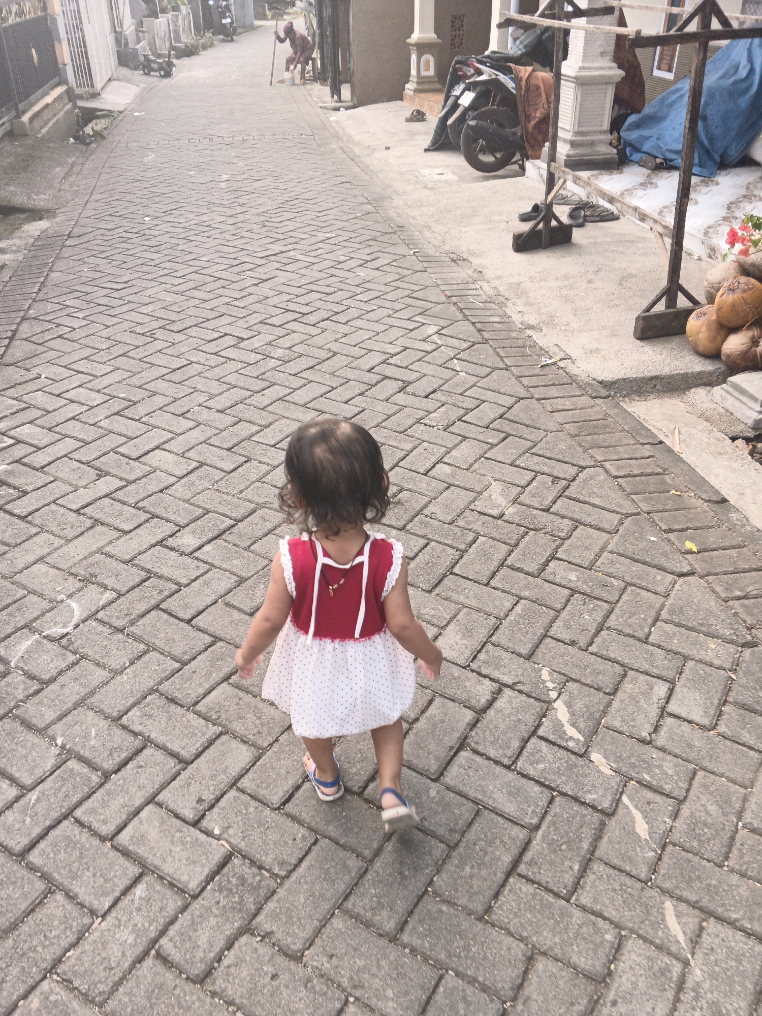 An elevated rear-view shot captures a toddler in a red and white polka dot dress walking down a herringbone-patterned brick pathway in Singolatren, Indonesia. The toddler is wearing blue and white sandals and has dark, curly hair. In the background, a person dressed in brown is stooping near a white bucket, holding a stick, adjacent to a building with white pillars. Further down the pathway, a motorcycle is parked. To the right of the pathway, a wooden stand displays coconuts and pink flowers. The scene appears to be a residential street with simple architecture.