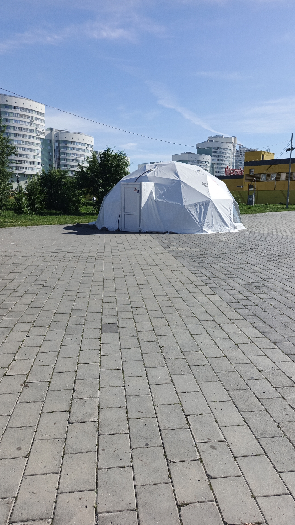 A geodesic dome tent, white in color and appearing to be made of fabric, stands in an open, paved area. The tent has a door with a white frame. Behind the tent, a line of green trees is visible, with several multi-story apartment buildings and a yellow building in the background. The sky is blue with some wispy clouds. The paved area is composed of rectangular grey stones. The scene appears to be located in an urban setting, likely in the city of Yekaterinburg, Russia, given the architectural style of the buildings. There are no people visible in the image.