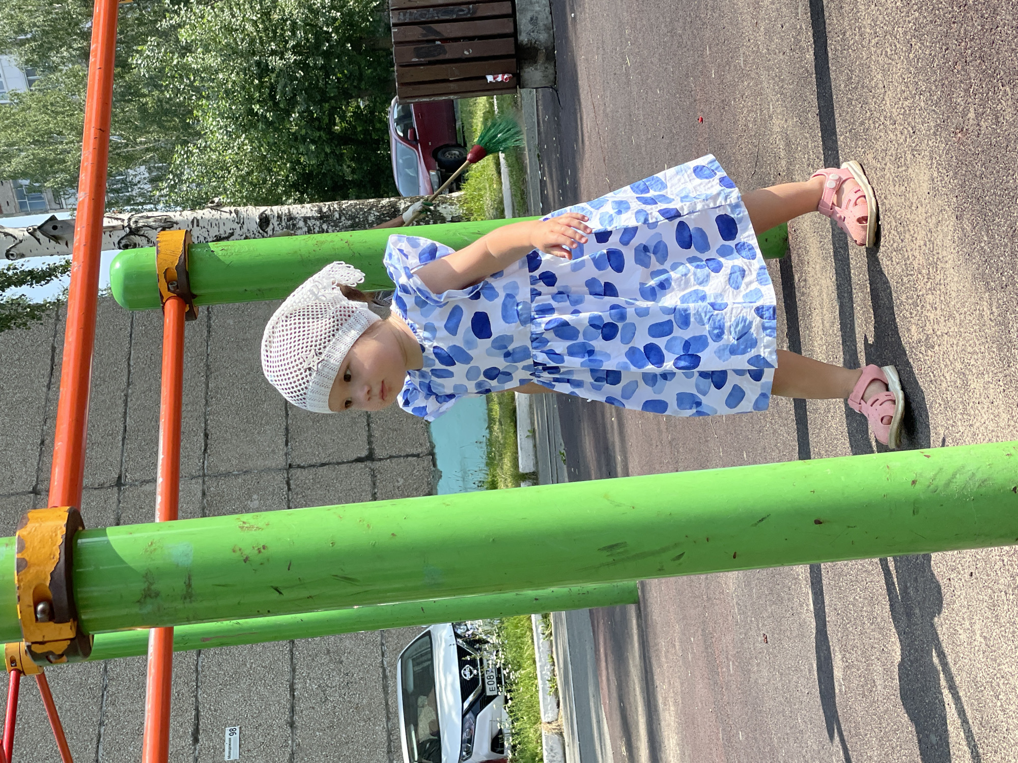 A young girl in a white and blue polka dot dress and a white head covering stands on a playground in Izhevsk, Russia. She is wearing pink sandals and looking to her right. The playground equipment consists of bright green poles and orange bars. In the background, a white car with license plate E087EE is parked near a building with a gray facade. To the right of the girl, a wooden trash bin and a red car are visible, with a broom leaning against the trash bin. Trees and foliage surround the playground.