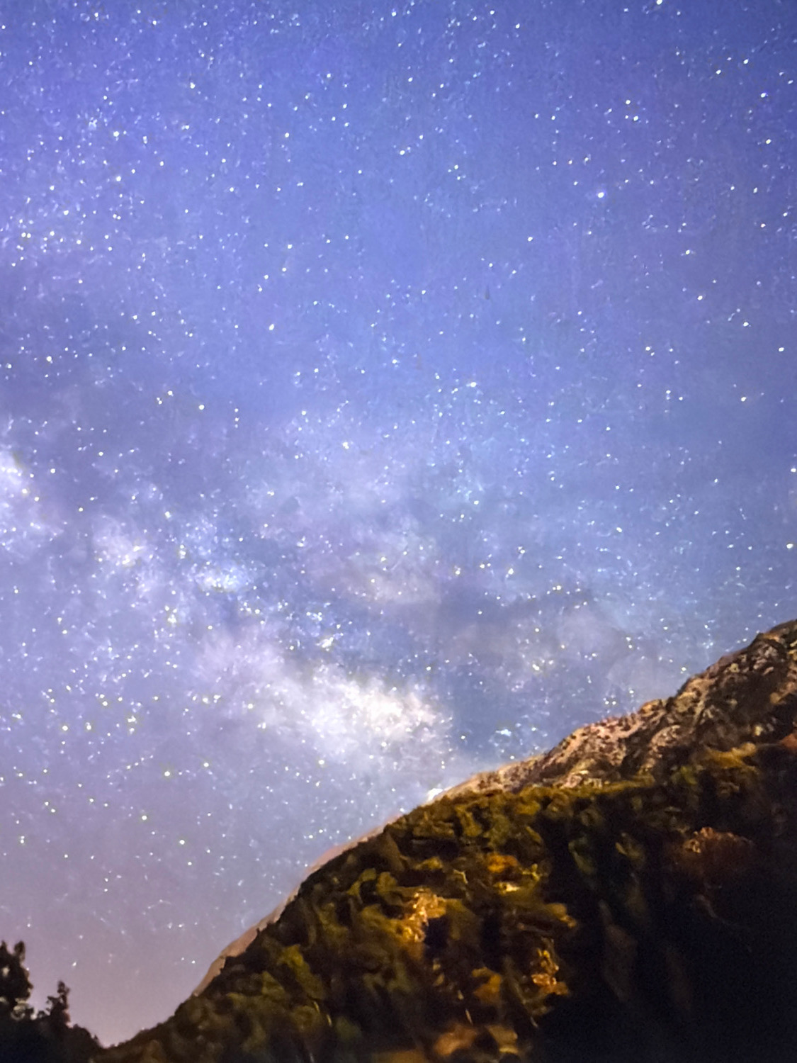 The image shows a starry night sky with the Milky Way visible as a band of light. A mountain range covered in trees is in the foreground, obscuring the lower portion of the sky. The stars are numerous and appear as small white dots against a deep blue and purple background. The Milky Way is a hazy, luminous band stretching across the sky, with clusters of brighter stars within it. The mountain is dark and rugged, with green vegetation covering its slopes. The lighting suggests a natural light source, likely moonlight or starlight, illuminating the scene. No people or man-made objects are visible. The image is captured from a location with minimal light pollution, allowing for a clear view of the night sky.