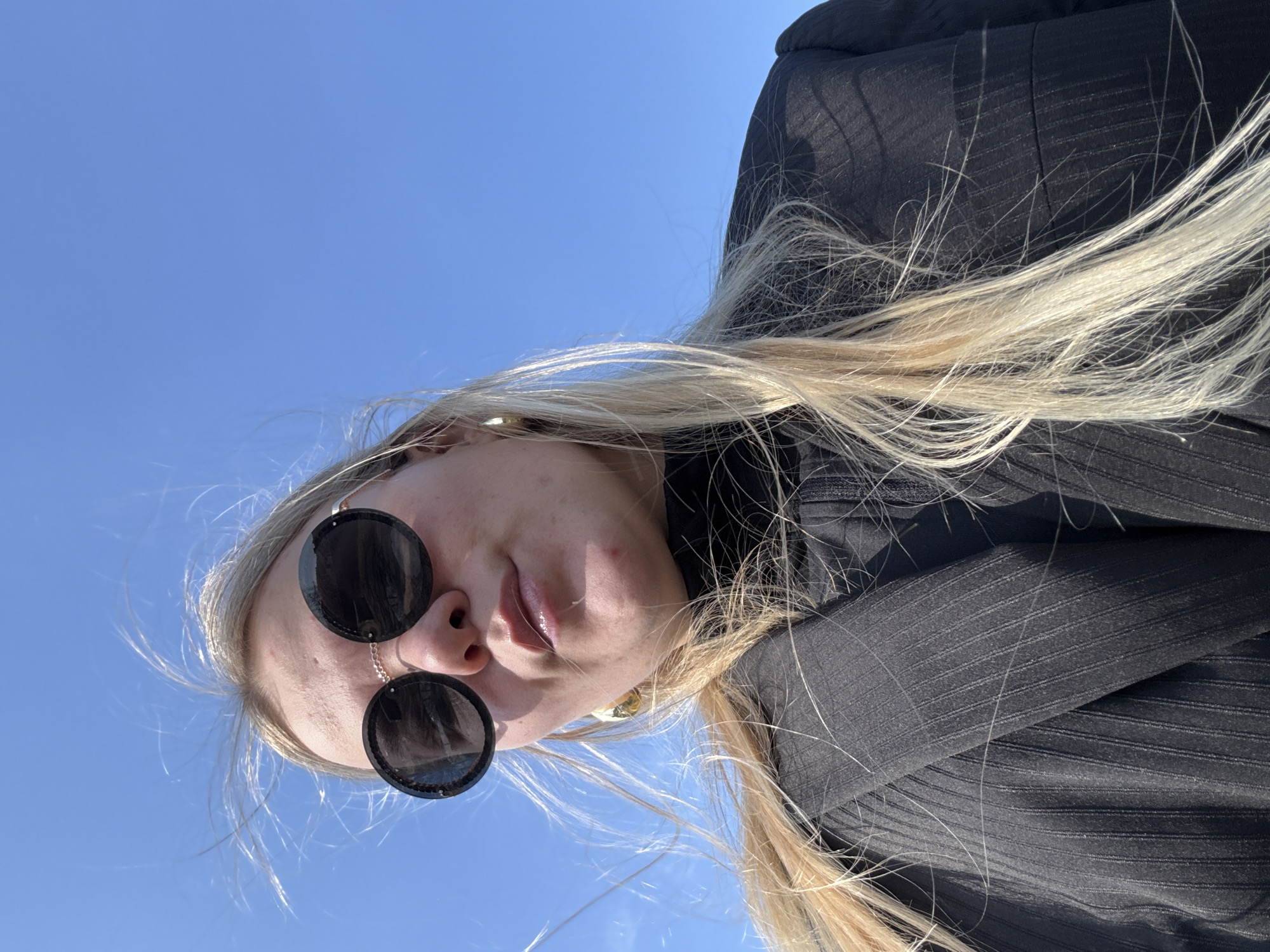 A close-up, low-angle photograph captures a person with long, flowing blonde hair and sunglasses set against a clear blue sky. The subject is oriented as if looking down at the camera, giving an inverted perspective. They wear large, dark, circular sunglasses, with a thin gold chain visible near the bridge of the nose, possibly part of the eyewear or an earring. Their lips are slightly parted, and their fair skin is visible around the sunglasses.
The person is dressed in a black garment, which appears to be a jacket or coat, characterized by a subtle ribbed or finely striped texture. Their long, light blonde hair is prominent, framing the face and cascading across the black clothing.
The background is an expansive, vibrant, and cloudless blue sky, indicating bright, sunny daytime weather conditions. The natural light highlights the blonde hair and the details of the black garment. The image conveys a casual outdoor moment, possibly a selfie taken from an unusual angle. The location context provided is Chaikovsky, Russia, though no specific geographical landmarks are visible in the frame. There is no visible text present in the image.