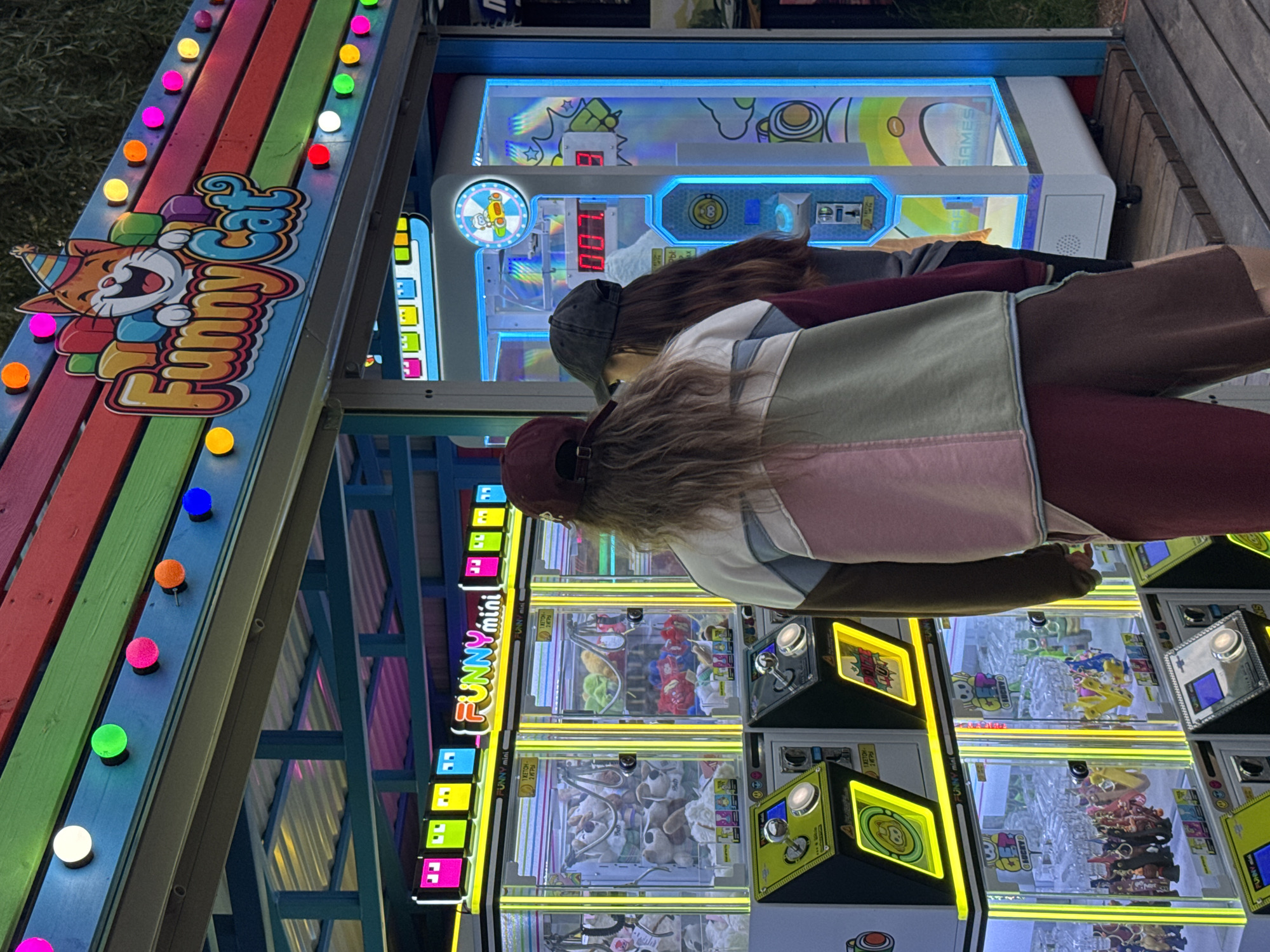 The image shows an outdoor scene at what appears to be a carnival or amusement park at dusk. In the foreground, there are several arcade claw machines, lit up with bright yellow and blue lights. The machines are filled with stuffed toys, including dogs and other characters. A sign above the machines reads "FUNNY mini".
Behind the claw machines, a colorful, striped structure with colorful lights is visible. This structure appears to be part of a game stall. There are also people in the image, seen from behind, observing or playing the games. One person is wearing a grey baseball cap and a maroon hoodie, while another has long brown hair and wears a pink and grey jacket.
The overall impression is one of a lively entertainment area at an evening event. The lighting suggests it is either early evening or has recently become dark. The presence of arcade games and bright lights evokes a sense of fun and entertainment.