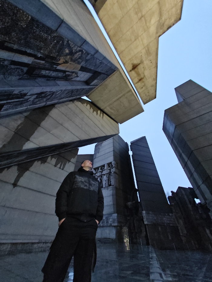 A low-angle, wide shot captures a single adult male standing within the Monument to 1300 Years of Bulgaria in Shumen, Bulgaria. The man, positioned in the lower-left foreground, wears a dark, padded jacket with a grey camouflage-patterned upper section, dark trousers, and dark footwear. His hands are in his pockets, and his head is tilted sharply upwards, directing his gaze towards the monumental concrete structures overhead.
The surrounding environment consists of massive, angular concrete slabs and walls characteristic of Brutalist architecture. Several large, light grey concrete beams cantilever outwards above, creating complex geometric forms and deep shadows. Portions of the monument's walls are adorned with dark, textured panels, and bas-reliefs depicting figures are discernible on vertical surfaces in the mid-ground, particularly to the right and behind the man. The ground surface directly beneath the subject is dark and partially reflective. A pale blue sky is visible between the imposing concrete elements at the top of the frame. The scene is illuminated by soft, ambient light, suggesting either dawn or dusk conditions.