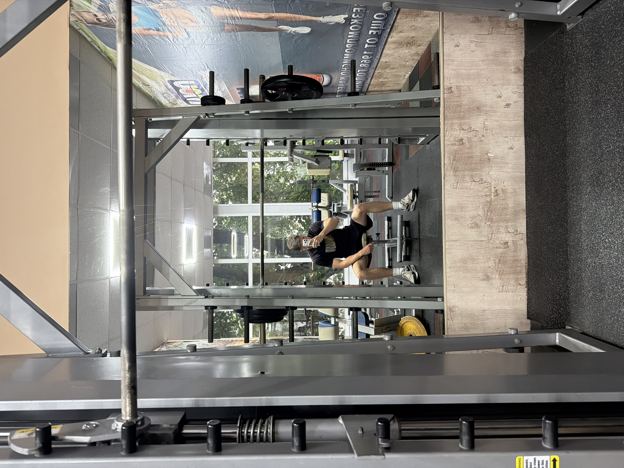 The image captures an indoor gym setting, viewed from a slightly elevated perspective looking down into the workout area. The scene is bright, indicating daytime, with ample natural light entering through large windows in the background that offer a view of green trees outside.
In the center of the frame, a male individual is visible, dressed in a black t-shirt, black shorts, and white sneakers. He appears to be positioned on or near a piece of gym equipment, possibly a squat rack or a similar weight training station. His right leg is bent with his foot on a raised surface, while his left leg is extended. He is holding a smartphone up with both hands, suggesting he might be taking a selfie, recording his workout, or checking his device. His face is largely obscured by the phone.
The gym is equipped with various metal structures, weight plates, and other exercise machinery. Grey and black floor tiles are visible on the right side, along with a section of light-colored wooden flooring. The foreground and sides of the image are framed by metallic parts of gym equipment, creating a structured, industrial feel.
A large banner or poster hangs on the wall or ceiling in the upper part of the image, featuring a person and some Cyrillic text. Due to mirroring and partial obstruction, parts of the text are difficult to read, but phrases like "ОТЛОЖИ" (Postpone) and "ЖИЗНЬ НА ПОТОМ" (Life for later) are discernible, likely part of an motivational slogan. A small, unreadable yellow sticker with safety instructions is visible on equipment in the lower left.
