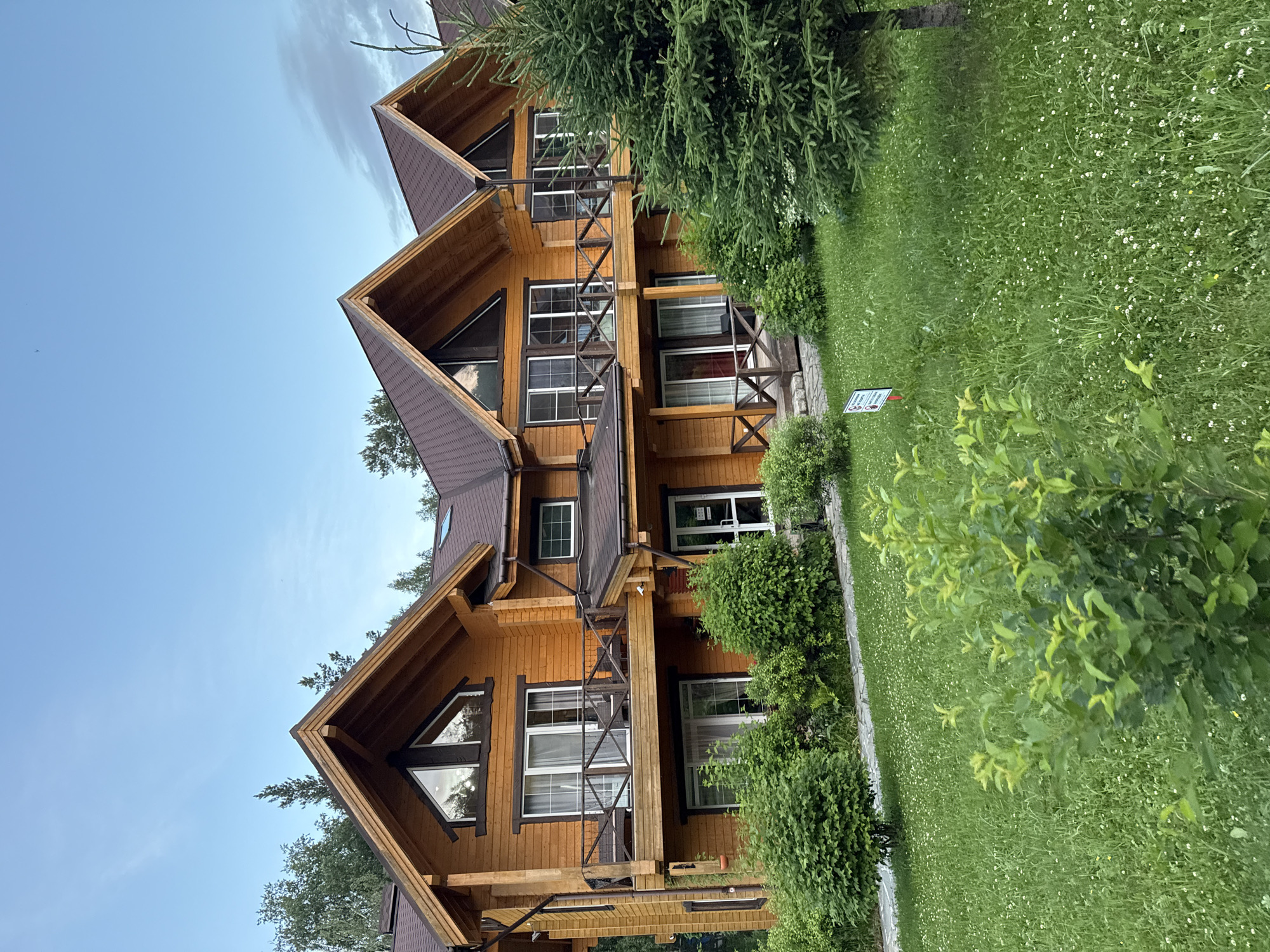 In Syrostan, Russia, a row of three identical wooden duplex houses stands on a slight incline covered in lush green grass dotted with small white flowers. The houses, constructed from light-colored wood, feature prominent gable roofs with dark brown shingles. Each unit has two stories, with the upper floors boasting balconies with wooden railings. Large windows and sliding glass doors are visible on both levels, offering views of the surrounding greenery. A large evergreen tree is positioned to the right of the houses, partially obscuring the rightmost unit. The sky above is a clear blue, with a few wispy clouds. The scene appears to be a tranquil residential or resort area.