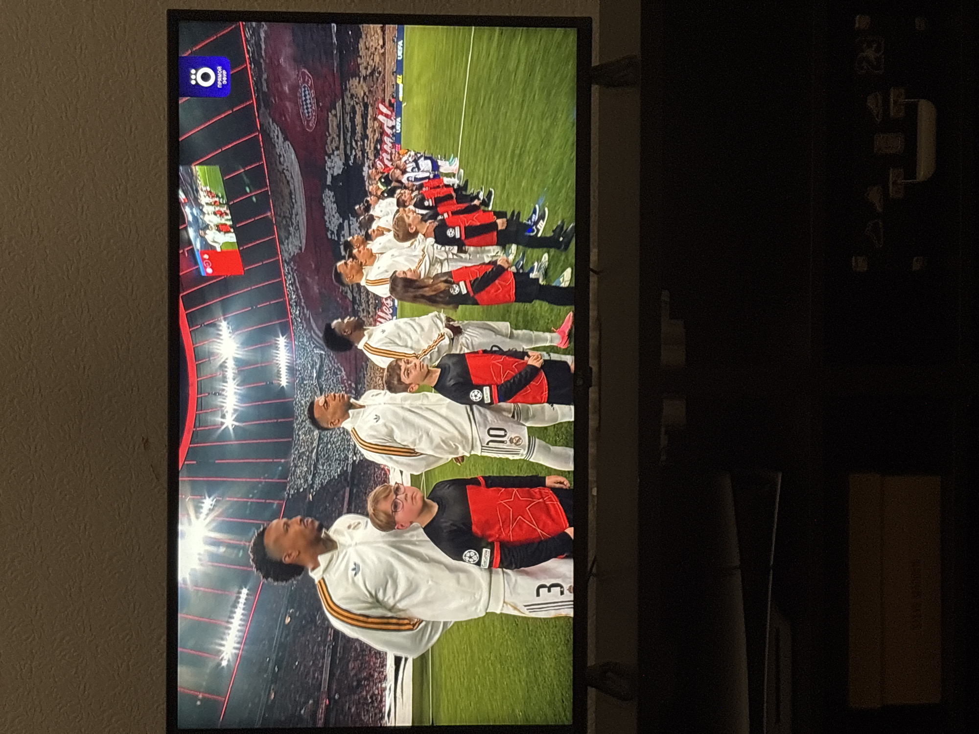The image shows a television screen displaying a soccer match. Players line up on a green field, wearing white and red jerseys. The background includes a stadium filled with spectators. The broadcast includes a logo in the upper left corner. The location is not discernible from this image.