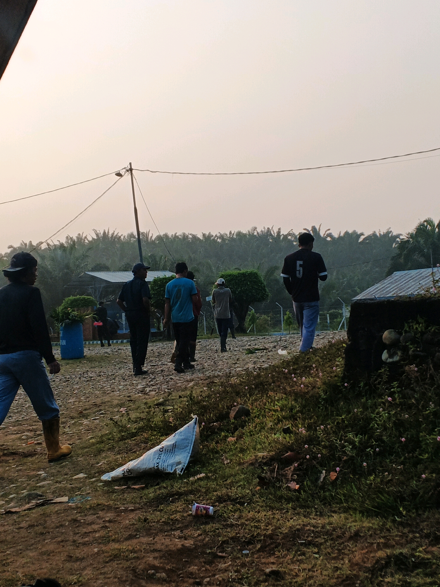 In Batu Ejung, Indonesia, a group of six individuals are observed walking on a gravel path adjacent to a dense palm tree plantation. The sky is overcast with a soft, diffused light, suggesting early morning or late afternoon. In the foreground, a white plastic bag and a discarded beverage can lie on the grassy ground. One individual, wearing a black shirt with the number "5" on the back, is walking away from the camera. Another person, in a blue shirt, is also walking away. To the left, a person in a black cap and blue jeans, wearing yellow boots, is seen walking toward the right. The environment appears to be rural, with a power line stretching across the frame, supported by a wooden pole. A blue barrel and some bushes are visible in the middle ground. A small, angled building with a metallic roof is situated to the right.