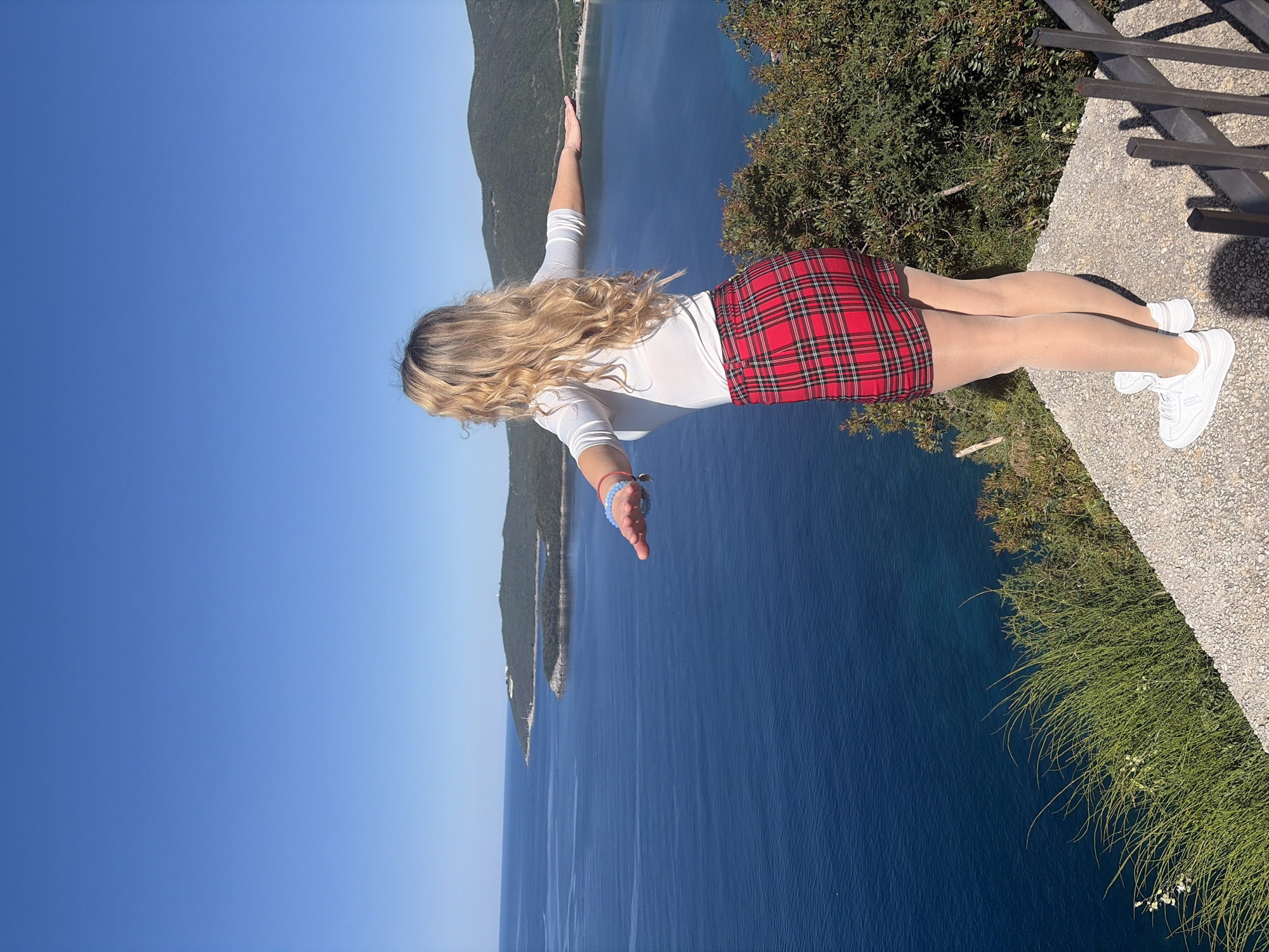 A woman is photographed outdoors. She is wearing a long-sleeved white shirt, a red plaid mini-skirt, nude-colored tights, and white sneakers. Her arms are outstretched, and she appears to be standing on a ledge.
The background consists of a blue body of water, a strip of land, and a clear blue sky. The water is a deep shade of blue, and the land is covered in green vegetation.
The photograph appears to be taken from a high vantage point and is rotated ninety degrees to the right. The location is not visible, and therefore the city of Budva, Montenegro, cannot be confirmed. A low fence or railing is visible on the right side of the image, along with greenery. A concrete ledge is also visible.