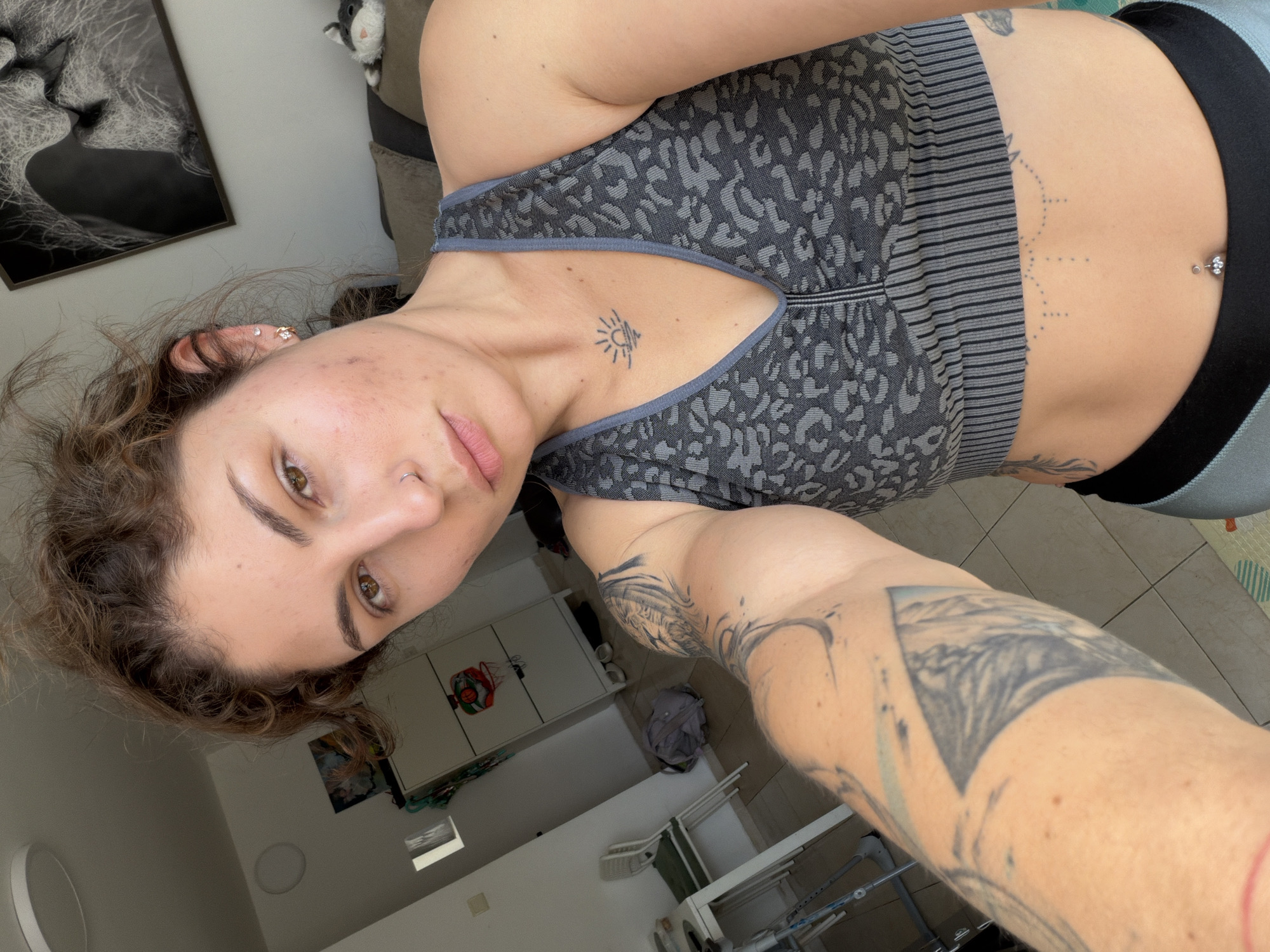 A young woman is posing for a selfie indoors. She has curly brown hair, fair skin, and brown eyes. She is wearing a gray patterned sports bra and light blue leggings with a black waistband. The woman has multiple tattoos including one on her chest, abdomen, and arm. In the background, there is a framed picture of two lions, a stuffed animal, and a white cabinet. The room appears to be a bathroom or utility room. There are other objects such as a drying rack, clothes, and a window. It is impossible to determine the exact location of the image, including within Kiryat Bialik, Israel.
