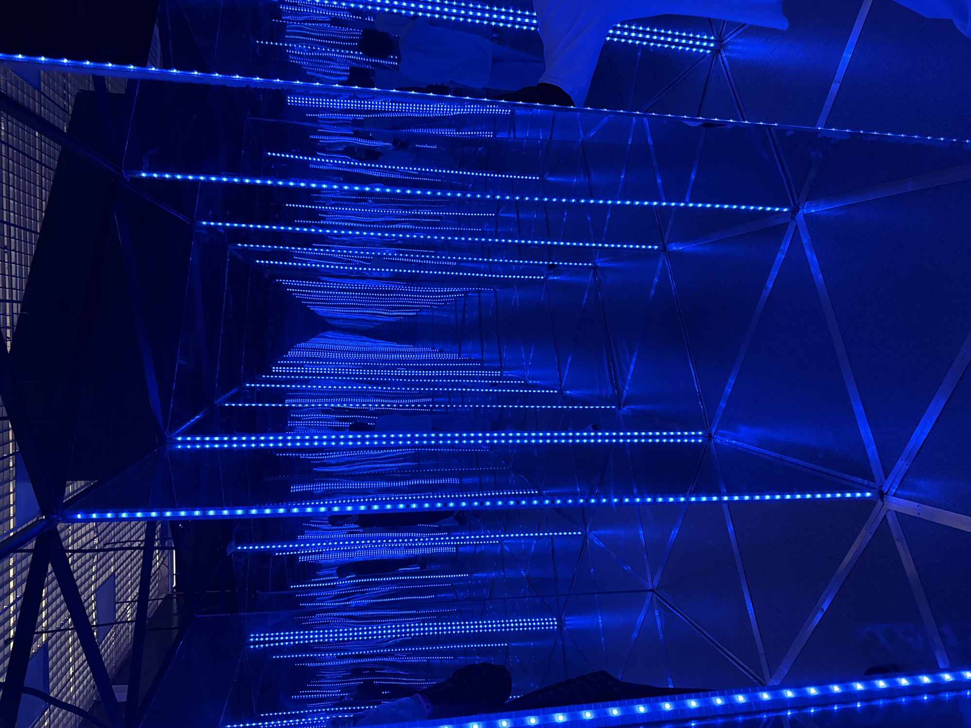 This media file captures an immersive art installation, likely an "infinity room" or similar exhibit, located in Moscow, Russia. The scene is dominated by electric blue LED light strips running horizontally, which are reflected repeatedly by mirrored surfaces on multiple planes, creating an illusion of an infinite, glowing tunnel.
The primary objects are the blue LED light strips, which are arranged in parallel lines and appear to stretch into the distance due to the reflections. These lights illuminate the dark environment, creating a high contrast scene. The surfaces creating the reflections are highly polished, likely mirrors or reflective panels, which form the walls, floor, and ceiling of the installation. Geometric structural elements are visible, particularly on the right side of the image, suggesting a faceted or geodesic design for the enclosure. On the far left, a distinct light-colored grid-like pattern is visible, possibly part of an external wall or another structural element.
Parts of at least one person are visible within the installation. In one area, a person's lower legs and feet, clad in white clothing, are seen from above, standing on a reflective surface. In another section, what appears to be a person's feet or lower body is visible at the bottom of the frame, also on a reflective surface. Their presence suggests they are observing or moving through the exhibit.
The overall atmosphere is futuristic and mysterious, with the vibrant blue lights creating a sense of depth and endlessness. The artificial lighting indicates an indoor setting, making the time of day and weather irrelevant. There is no visible text in the image. The installation evokes a sense of wonder and visual disorientation, typical of such immersive art experiences.