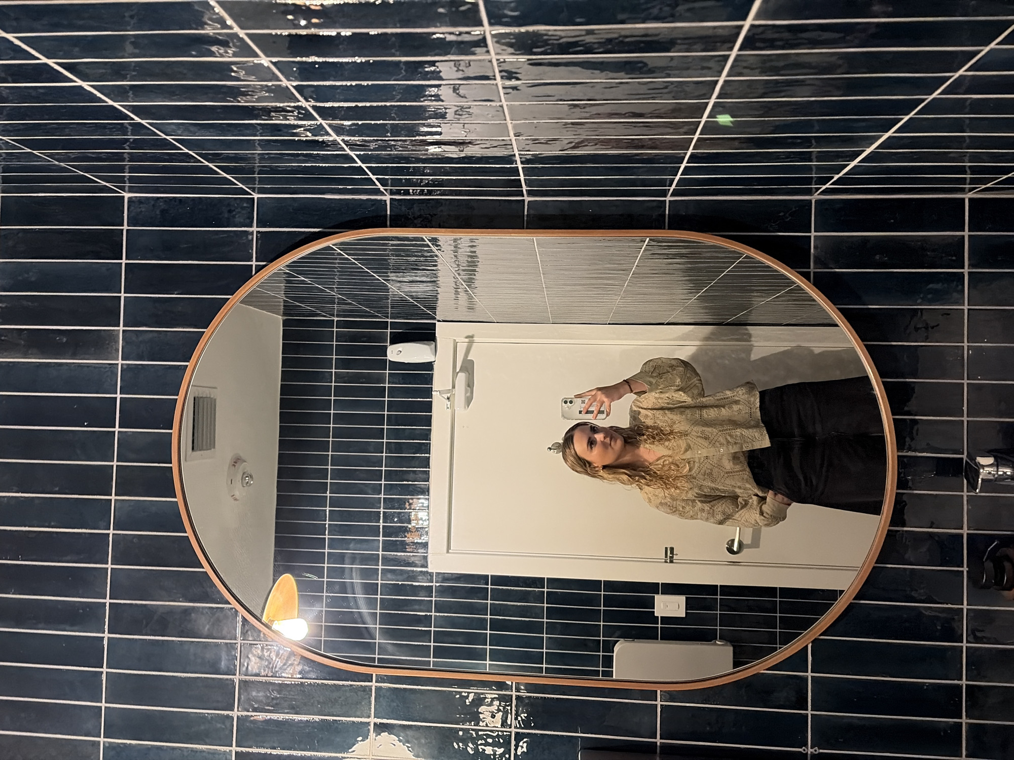 A mirror selfie captures a woman in what appears to be a modern bathroom or restroom, located in Coral Gables, United States of America. The walls are covered in striking dark blue, glossy rectangular tiles with clean white grout lines, providing a sleek and textured backdrop. An oval-shaped mirror with a thin, light brown wooden frame is central to the composition.
Reflected in the mirror, a woman with long, curly blonde hair is seen taking the photo. She is wearing a light-colored, possibly beige or off-white, long-sleeved top with a subtle pattern and dark pants or a skirt. She holds a white smartphone with her right hand to capture the image, looking directly at the camera with a neutral expression.
The reflection also reveals details of the room: a white door with a silver handle and a latch is visible behind the woman. To the left of the reflected door, a white wall-mounted dispenser (possibly soap or hand sanitizer) is affixed to the tiled wall. Inside the reflected room, above the door, a white ceiling vent and a circular fire safety device (like a smoke detector or sprinkler) can be seen. To the right of the woman's reflection, part of a white toilet tank or a small counter and some metallic fixtures are partially visible. The space appears well-lit, highlighting the reflective surfaces. No distinct text is visible in the image.