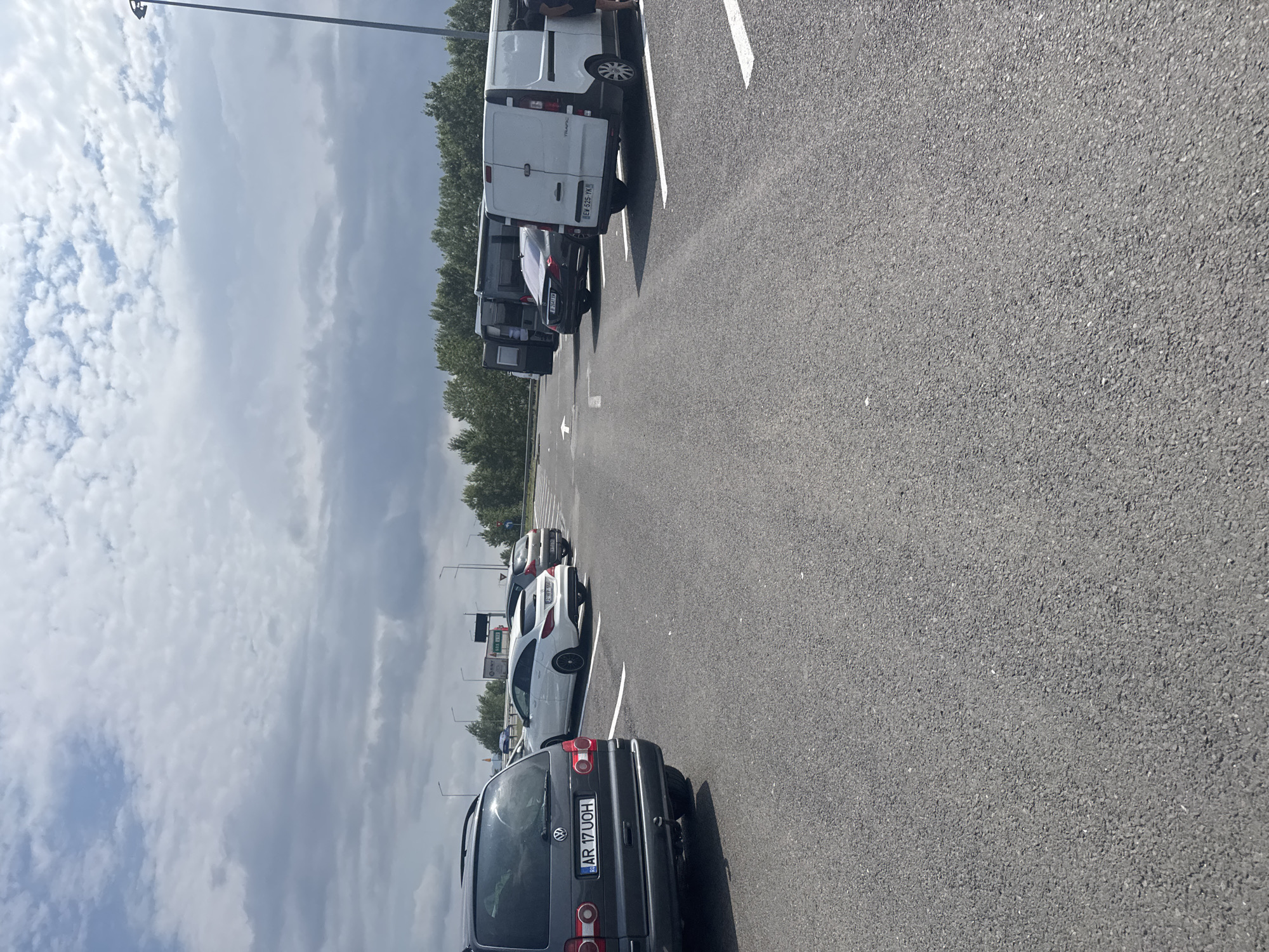 A parking lot at a service area along a highway outside of Orăștie, Romania, features several vehicles. In the foreground, a dark gray Volkswagen Sharan minivan is parked on asphalt. White parking lines mark the spaces. To the right of the minivan, a white Mercedes-Benz CLA station wagon is parked, followed by a dark-colored sedan and a white van. Several other vehicles, including a dark van and a lighter van, are parked further back. The environment is an open, paved area with sparse trees and vegetation in the background. The sky is partly cloudy with a mix of white and gray clouds. Streetlights are visible along the edge of the parking lot.