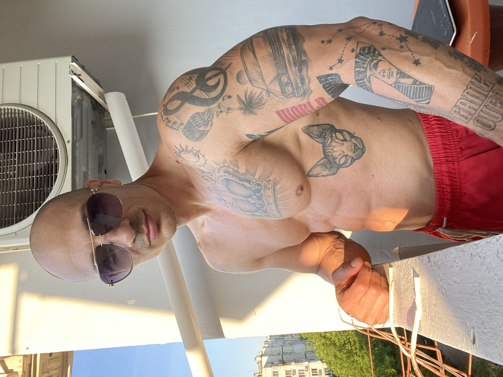 A man with a shaved head and sunglasses stands on a balcony, his torso bare, revealing extensive tattoos on his chest and arms. He wears red shorts. The man's body is muscular, with a defined six-pack. Tattoos visible include a crown on his chest, a cat's head on his abdomen, musical notes, a microphone, a classic car, a pharaoh's face within a number 7, and text that appears to read "GAME OVER" on his arm. Behind him, an air conditioning unit is mounted on the wall. The balcony railing is visible in the foreground, and beyond it, buildings and trees suggest an urban setting. The lighting indicates it is likely late afternoon or early evening, with warm sunlight casting shadows. The specific location is a balcony in the city of Pleven, Bulgaria.