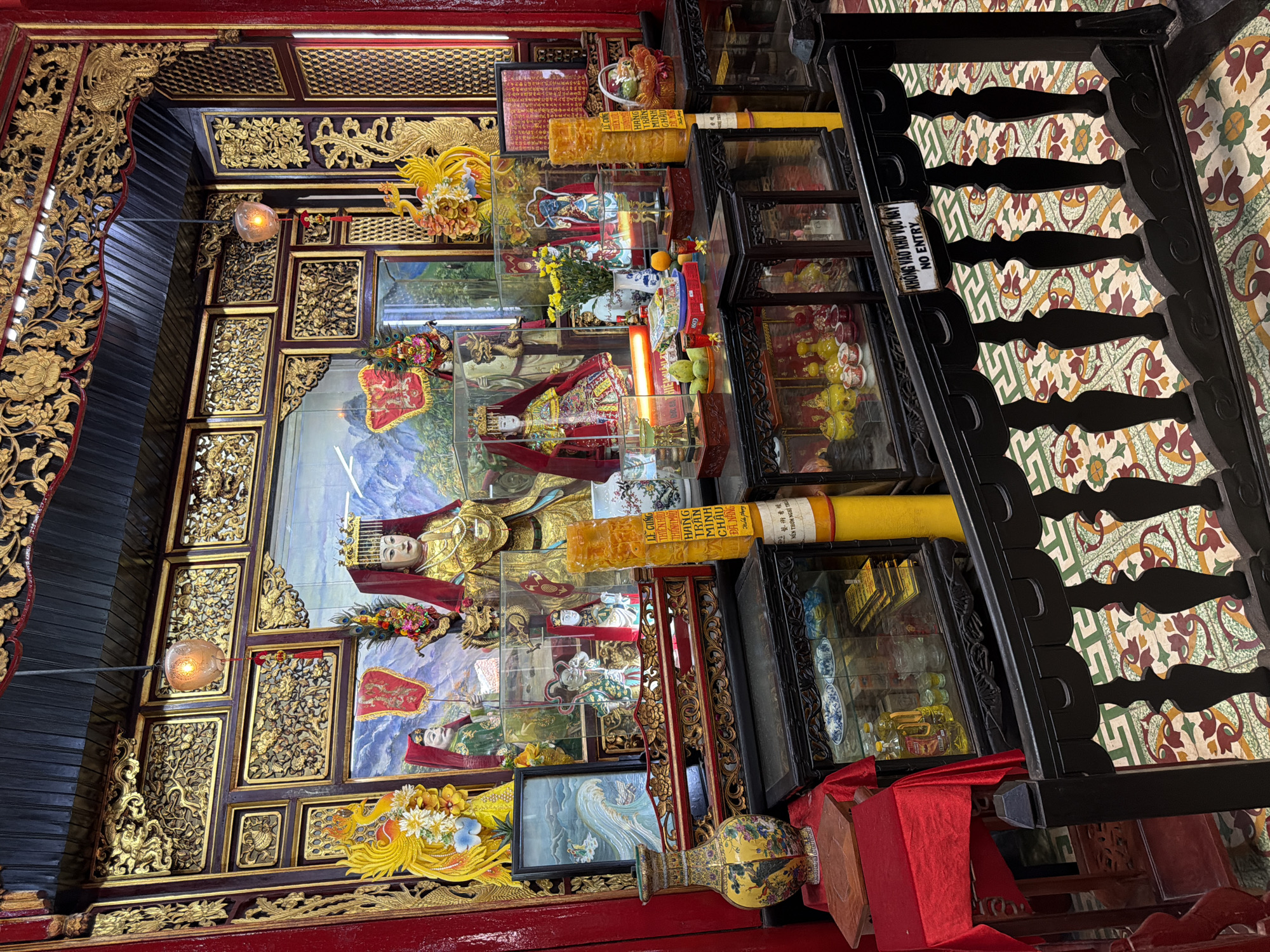 This image depicts an ornate altar within a temple in Hội An Ward, Vietnam. The altar is adorned with intricate golden carvings, religious statues, and offerings. Two prominent statues of figures in elaborate golden robes and headdresses are positioned centrally. To the left and right of these figures are smaller statues and decorative elements, including golden phoenixes. Vases, bottles of oil, fruit, and other offerings are displayed on tables and in glass cabinets. A painted backdrop featuring mountains and skies forms the rear of the altar. Yellow cylindrical candles, one with Vietnamese text, are prominent. A black wooden railing with decorative carvings separates the altar area from the viewer, with a sign reading "KHÔNG VÀO KHU VỰC NÀY NO ENTRY" attached to it. The floor is tiled with a colorful, patterned mosaic. The overall impression is one of religious devotion and cultural richness.