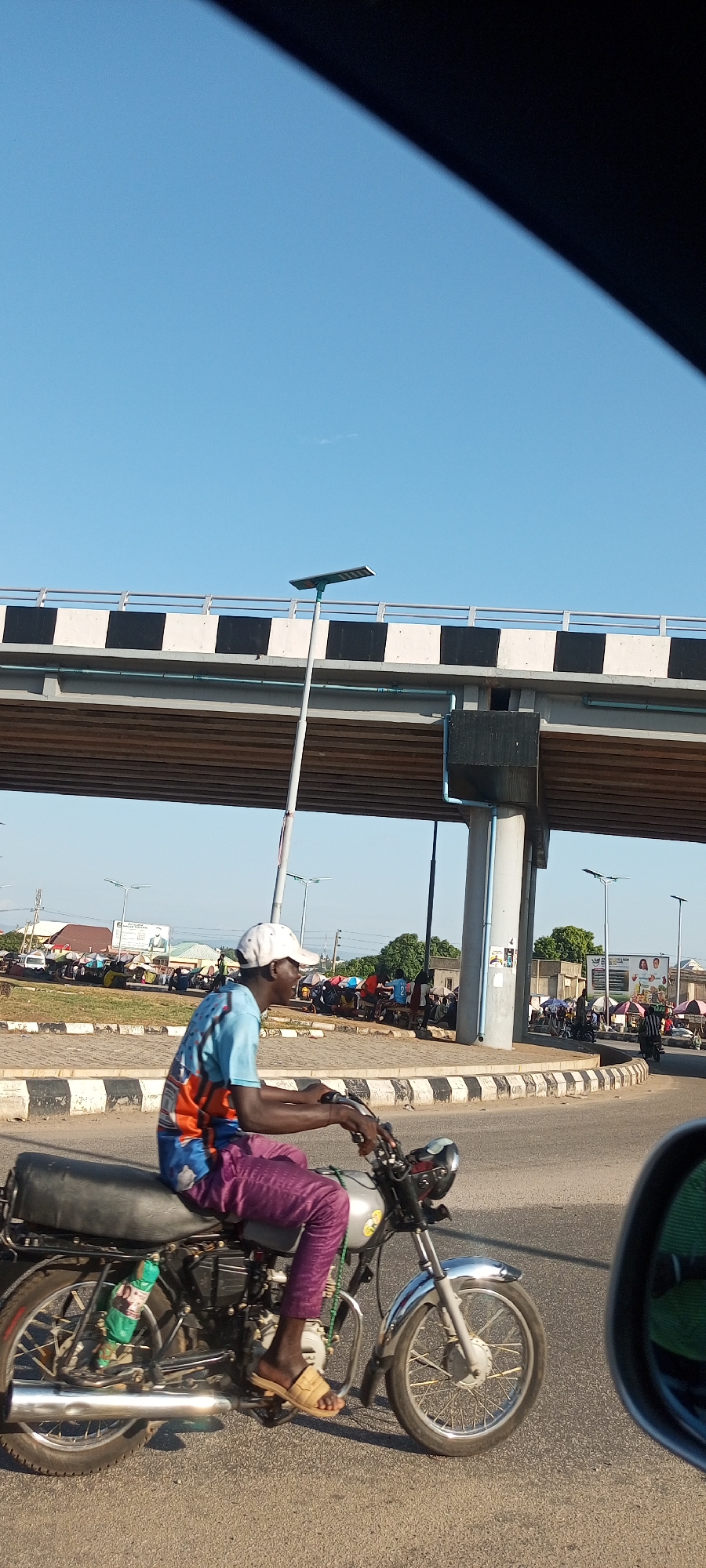 The image captures a vibrant street scene in Lokoja, Nigeria, under a clear, bright blue sky. In the foreground, a man with dark skin, wearing a white baseball cap, a light blue and orange patterned t-shirt, and purple patterned trousers, rides a silver motorcycle. He is wearing light-colored sandals and has a slight smile on his face, looking towards the right side of the frame. The motorcycle has a black seat and a green object, possibly a bottle, wrapped and attached near the rear wheel. A car's side mirror is visible in the bottom right, indicating the photo was taken from within a vehicle.
Dominating the upper half of the background is an elevated road or bridge with a distinct black and white striped barrier and metal railings. Beneath the bridge, modern solar-powered street lights stand tall. Further in the background, a bustling market or collection of stalls with colorful canopies and umbrellas is visible, suggesting active commerce. Buildings and trees line the horizon. The road features black and white striped curbs, common in the region. The scene depicts a typical day in a bustling urban environment, with people engaged in daily transit and market activities. No specific text is clearly legible on any signs or billboards.