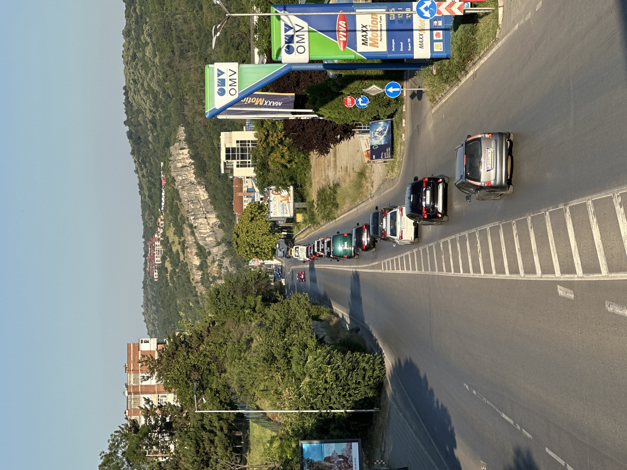 This image, taken in Veliko Tarnovo, Bulgaria, depicts a daytime scene of a road winding through a hilly landscape. The road is lined with trees and vegetation on one side, and a steep, rocky hillside with buildings on the other. Several cars are driving on the road, some appearing to be in a traffic jam near a gas station.
The gas station is a prominent feature, with OMV branding visible on its signs, along with "VIVA MAXX Motion Performance Fuels." Text on the gas station signs indicates fuel prices such as "Autogas D.E5," "Diesel," and "Super 95." Road signs are also visible, including a "STOP" sign and a blue arrow indicating a direction.
In the background, more buildings can be seen nestled on the hillside. The time of day appears to be late afternoon, judging by the long shadows cast by the trees. The weather seems clear and sunny. The scene conveys a sense of everyday life and travel, with the gas station serving as a functional point along the route. The overall impression is that of a typical roadside view in a scenic, somewhat urbanized environment.