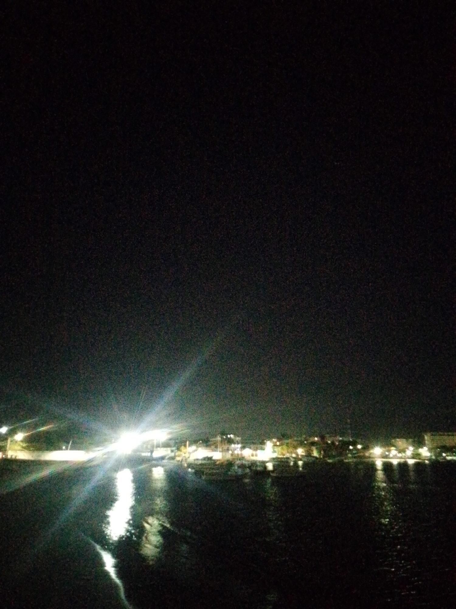 At night, a body of water is visible in the foreground, its surface reflecting the lights of the city of Juan Griego, Venezuela, located on the opposite shore. The light source on the left is particularly intense, obscuring details in that area. Multiple buildings are visible along the coastline, and other light sources are present along the shore. The sky is dark.