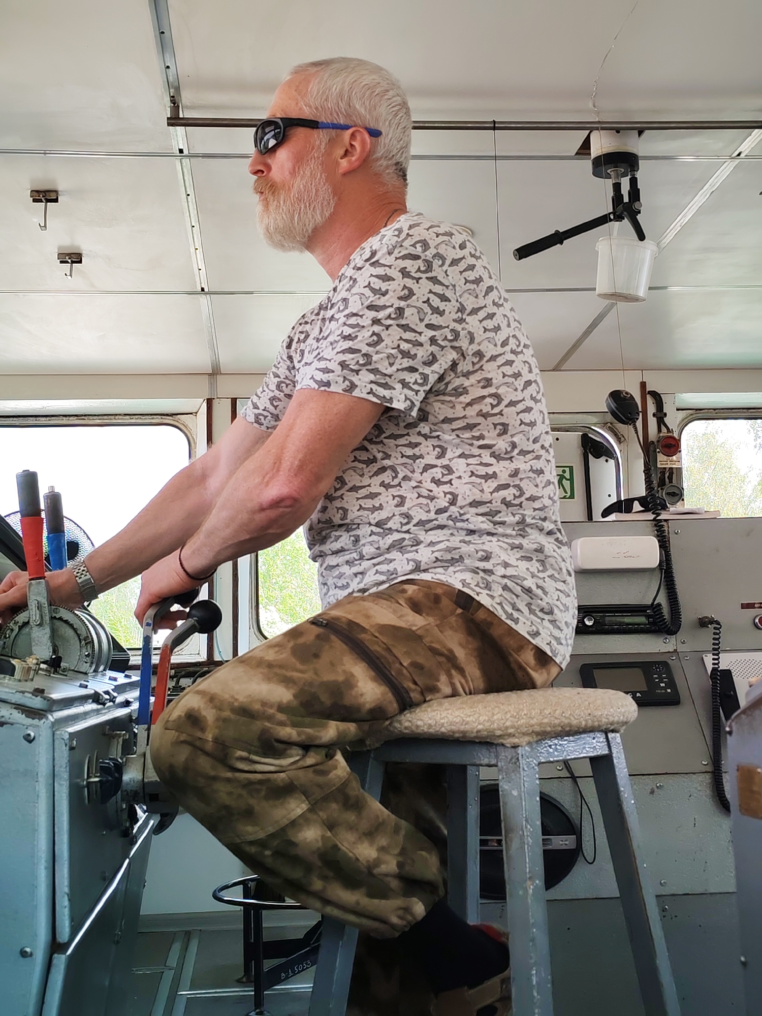 The image shows a man with white hair and a beard, wearing sunglasses, a t-shirt with a fish pattern, and camouflage pants, sitting on a padded stool inside a boat's control room. He is operating the boat's controls. The interior of the control room appears functional with metal surfaces and various equipment. The location is Unknown, Unknown.