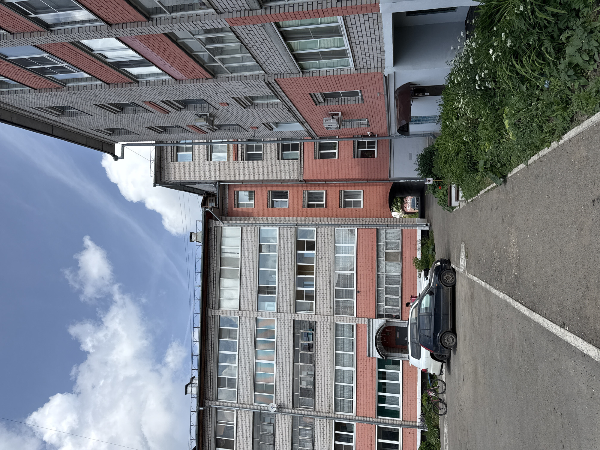 The image depicts a residential courtyard in Vologda, Russia, on a cloudy day. In the foreground, a paved area leads toward two apartment buildings. A black car is parked, and a bicycle is leaning against a white SUV behind it. The buildings are constructed with gray and pink bricks, featuring numerous windows and balconies. Lush green vegetation lines the side of the paved path, adding a touch of nature to the urban setting. The sky is partly cloudy, with fluffy white clouds scattered across a blue expanse.