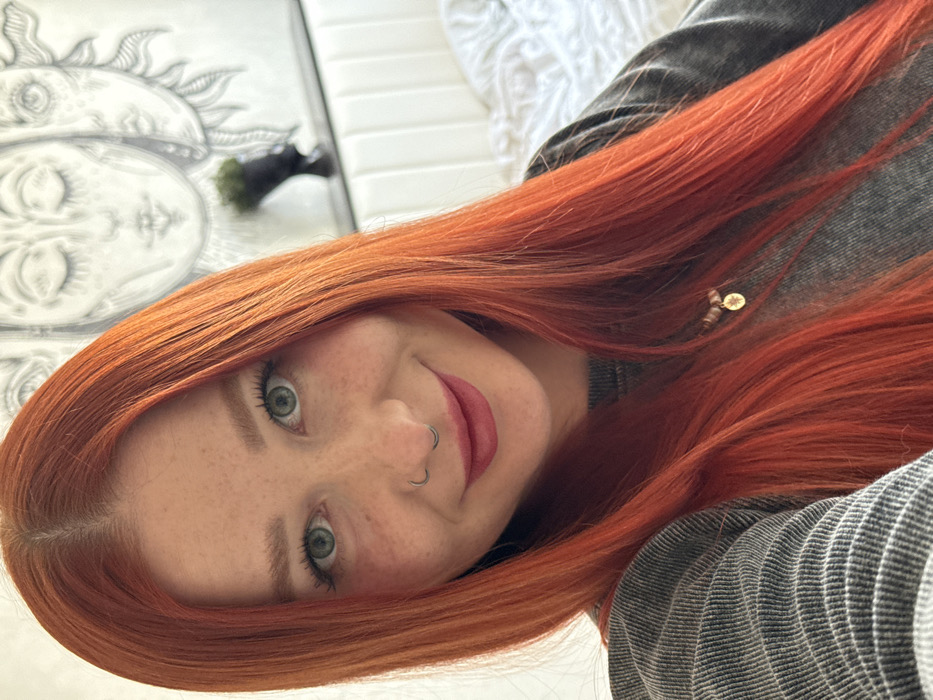 The image displays an indoor close-up portrait of a female individual in Rostov-on-Don, Russia. The subject, positioned in the center, has long, straight, dyed red-orange hair with a central part, a fair complexion with visible freckles across her nose and cheeks, and light-colored eyes. She wears a silver-colored circular septum ring and has dark pink lipstick applied. Her expression is a subtle smile directed at the camera. She is dressed in a grey, textured long-sleeved top, with a necklace featuring a small, gold-colored circular charm visible on her chest. The background suggests a residential bedroom environment, featuring a monochrome, celestial-themed wall art piece depicting multiple stylized faces behind her upper right side. Below the artwork, a white headboard with vertical paneling is partially visible, alongside a white bedspread. A dark, ornate vase containing dark foliage is situated on a shelf above the headboard.