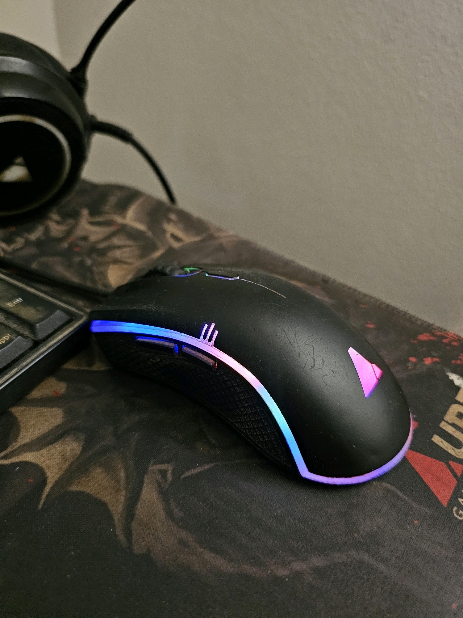 A black gaming mouse with RGB lighting is positioned on a black mousepad with a dark, abstract pattern. Part of a black keyboard is visible to the left of the mouse. In the background, a black headphone with a silver logo is partially visible. The lighting suggests an indoor environment, likely a desk setup. There is no indication of the city of Batna, Algeria, or any specific geographical markers.