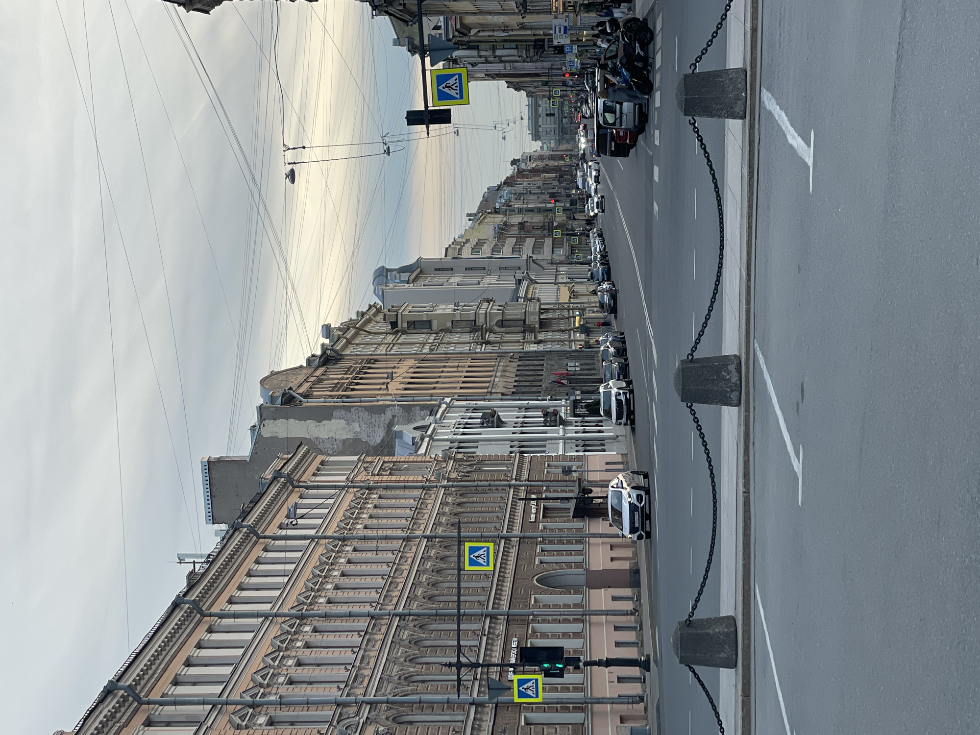 This detailed street scene depicts Liteyny Prospekt in Saint Petersburg, Russia, viewed looking south along the avenue under an overcast sky.
In the immediate foreground, a wide section of asphalt pavement shows faded white parking lines. Separating this area from the main thoroughfare is a concrete curb with a pedestrian barrier consisting of two dark gray, irregular stone bollards linked by a heavy black metal chain.
The midground features a multi-lane asphalt street with active vehicle traffic. On the left, a five-story building with a light pinkish-brown facade dominates, characterized by numerous tall windows, decorative molding, and vertical drainpipes. Signs on this building are in Cyrillic, including "Агент-Центр СПБ" (Agent-Center SPb), confirming its location in Saint Petersburg. A tall street pole stands near this building, displaying a green traffic light and two yellow square pedestrian crossing signs featuring a white walking figure. A white SUV, likely a Hyundai, with a Russian license plate, is visible driving away from the viewer. Further down the street on this side, several other buildings line the avenue, and a lone cyclist is visible.
On the right side of the street, a continuous row of historic multi-story buildings with varied facades (including white, gray, and brown tones) extends into the distance. Several cars are parked along the curb. Closer to the foreground, a dark minivan or SUV is parked, facing the viewer. Adjacent to it, a black scooter or motorcycle is parked, and several people are standing on the sidewalk, some near these vehicles. A second street pole with a red traffic light and a similar yellow pedestrian crossing sign is visible on this side.
The background shows Liteyny Prospekt receding into the distance, lined by more buildings and occupied by numerous vehicles, both moving and parked. Multiple overhead wires, likely for street lighting or public transport, crisscross the sky. The sky itself is uniformly gray and overcast, suggesting diffuse light without direct sunlight.