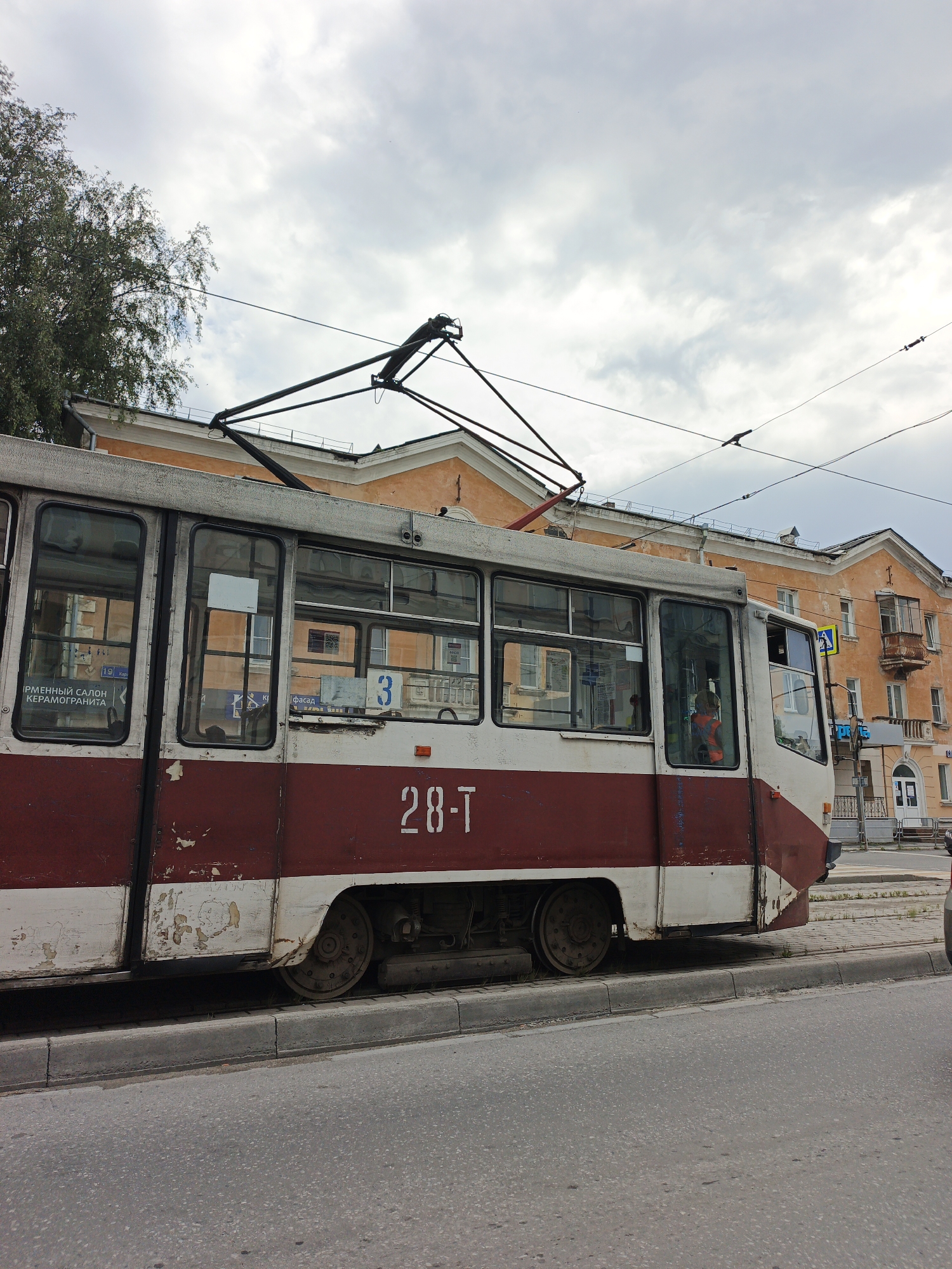 A worn, two-tone red and white tram, identified by the number "28-T" on its side, is parked at a curb on a street in Zlatoust, Russia. The tram, with its metal wheels and overhead electrical connection, shows signs of age, including peeling paint and rust. Visible through the tram's windows are reflections of buildings and a few indistinct passengers. Behind the tram, an orange-colored, multi-story building with a balcony and arched windows lines the street. The sky is overcast with clouds, and a large tree with dark foliage is present on the left side of the frame. The street is paved, and a concrete curb separates the tram's tracks from the roadway.