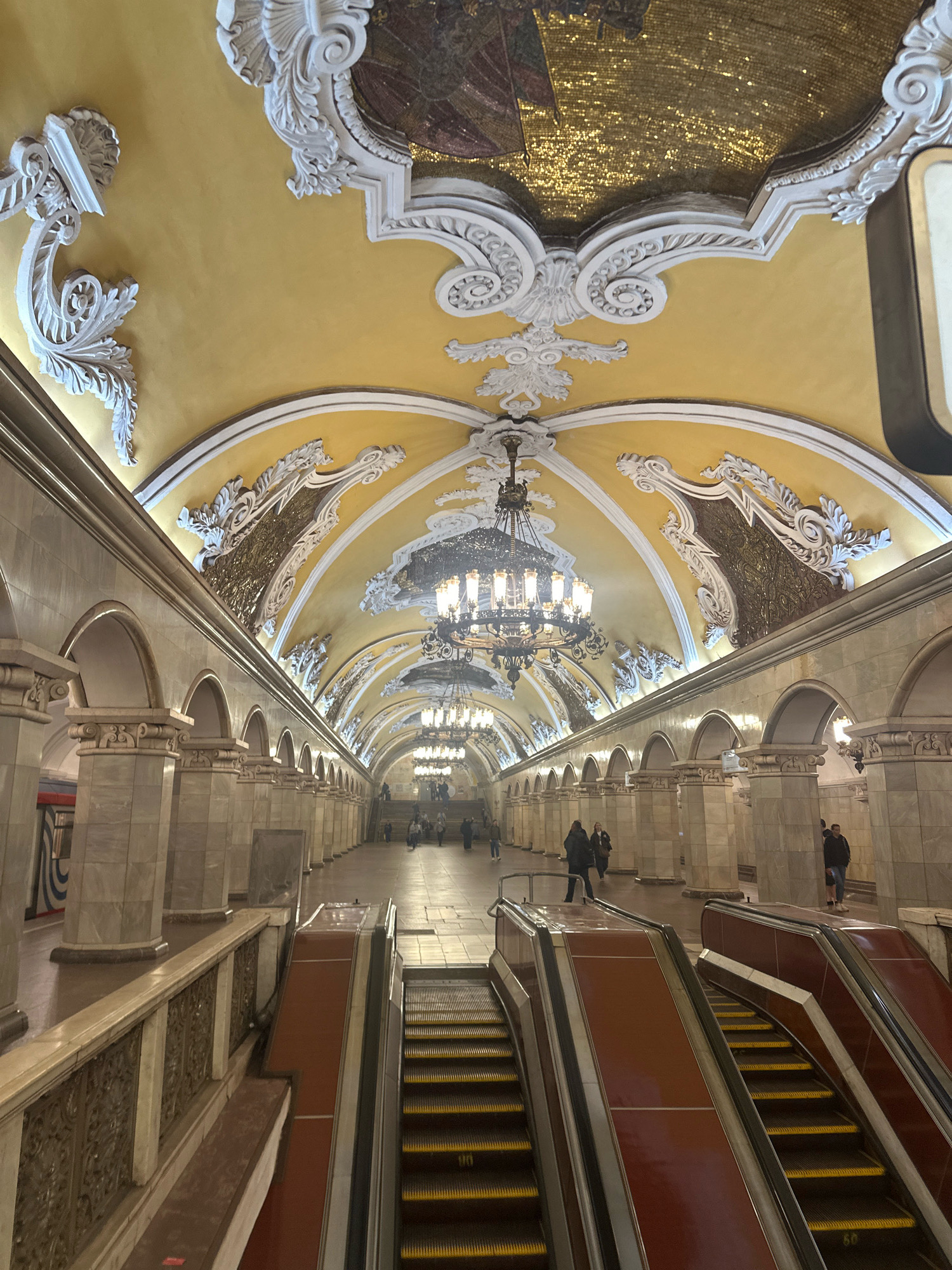 The image captures the ornate interior of a Moscow Metro station, likely one of the more grandly decorated ones. The dominant color scheme is a rich yellow on the vaulted ceiling, adorned with intricate white stucco moldings that frame mosaic panels. Large, elaborate chandeliers hang from the ceiling, illuminating the spacious hall.
The station features a long platform lined with imposing marble columns and arched openings, suggesting a classical or Baroque architectural style. In the foreground, a pair of escalators lead down into the station. The escalator treads are yellow, and the sides are a deep red-brown.
Several people are visible in the distance on the platform and at the top of the escalators, appearing to be passengers or commuters. They are dressed in casual clothing. The overall impression is one of grandeur and historical significance, typical of many Moscow Metro stations.