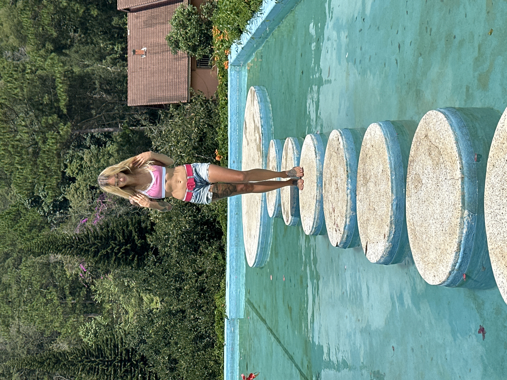 A young woman with blonde hair is walking on circular stepping stones across a pool of turquoise water. She is wearing a pink bikini top, denim shorts, and a pink belt. The stepping stones are textured and appear to be made of concrete, with a blue trim. The pool is surrounded by lush greenery and trees, with a rustic building visible in the background. The scene appears to be set in a natural, outdoor environment. The location is identified as Cam Ly - Đà Lạt, Vietnam.