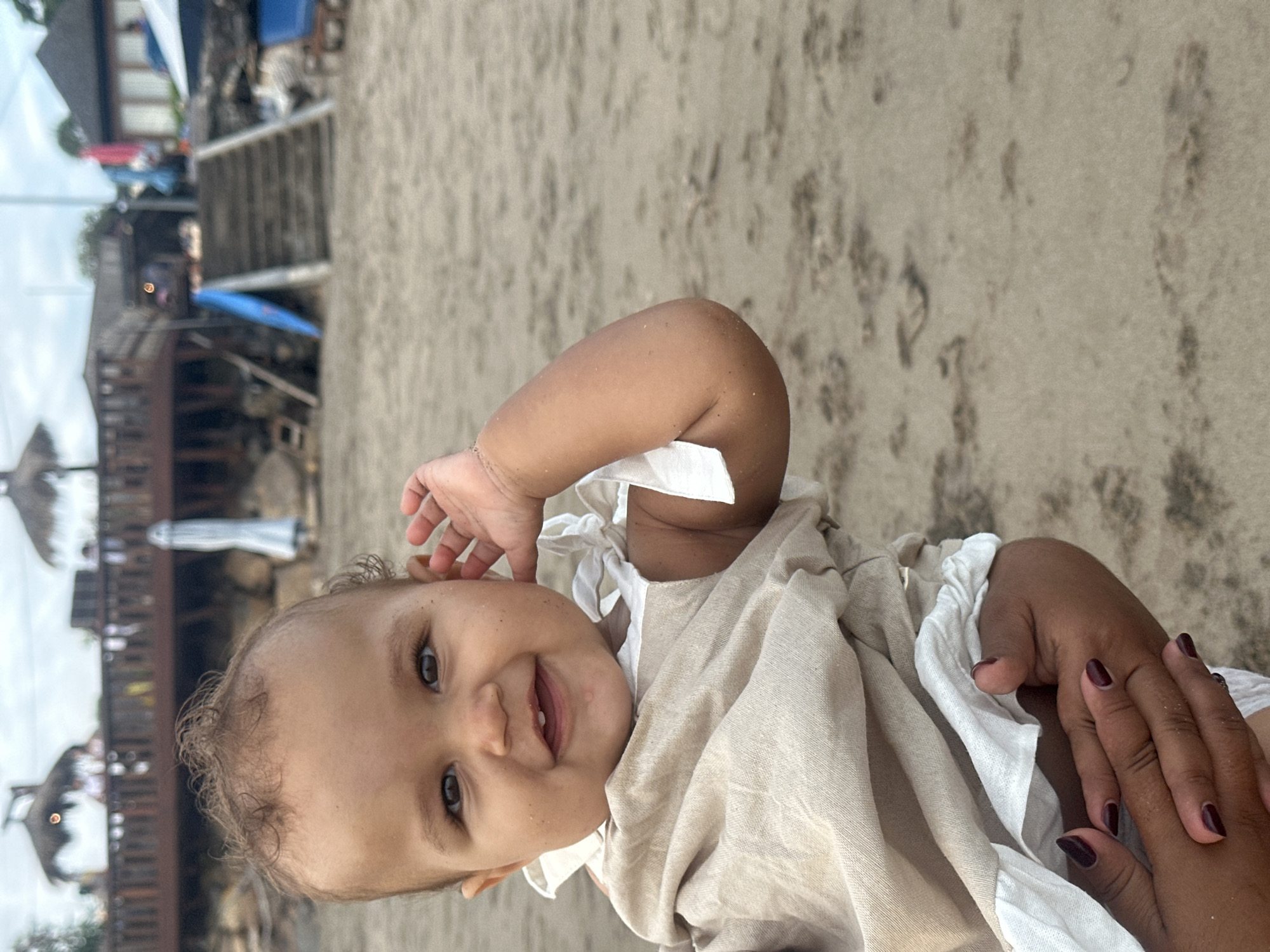 A close-up, over-the-shoulder shot shows a baby on a sandy beach in North Kuta, Indonesia. The baby, with dark, curly hair and large, dark eyes, is looking forward with a wide smile and two teeth visible. The baby is wearing a light tan romper with white ruffled trim. The baby's right hand is raised to their ear, and their left arm is resting on an adult's hands, which are visible at the bottom of the frame. The adult's hands have dark, manicured nails and are holding the baby's arm. The background shows a blurred view of a wooden deck with thatched-roof structures, stairs leading up to a building, and umbrellas on the beach. The overall lighting is soft, suggesting an overcast day.