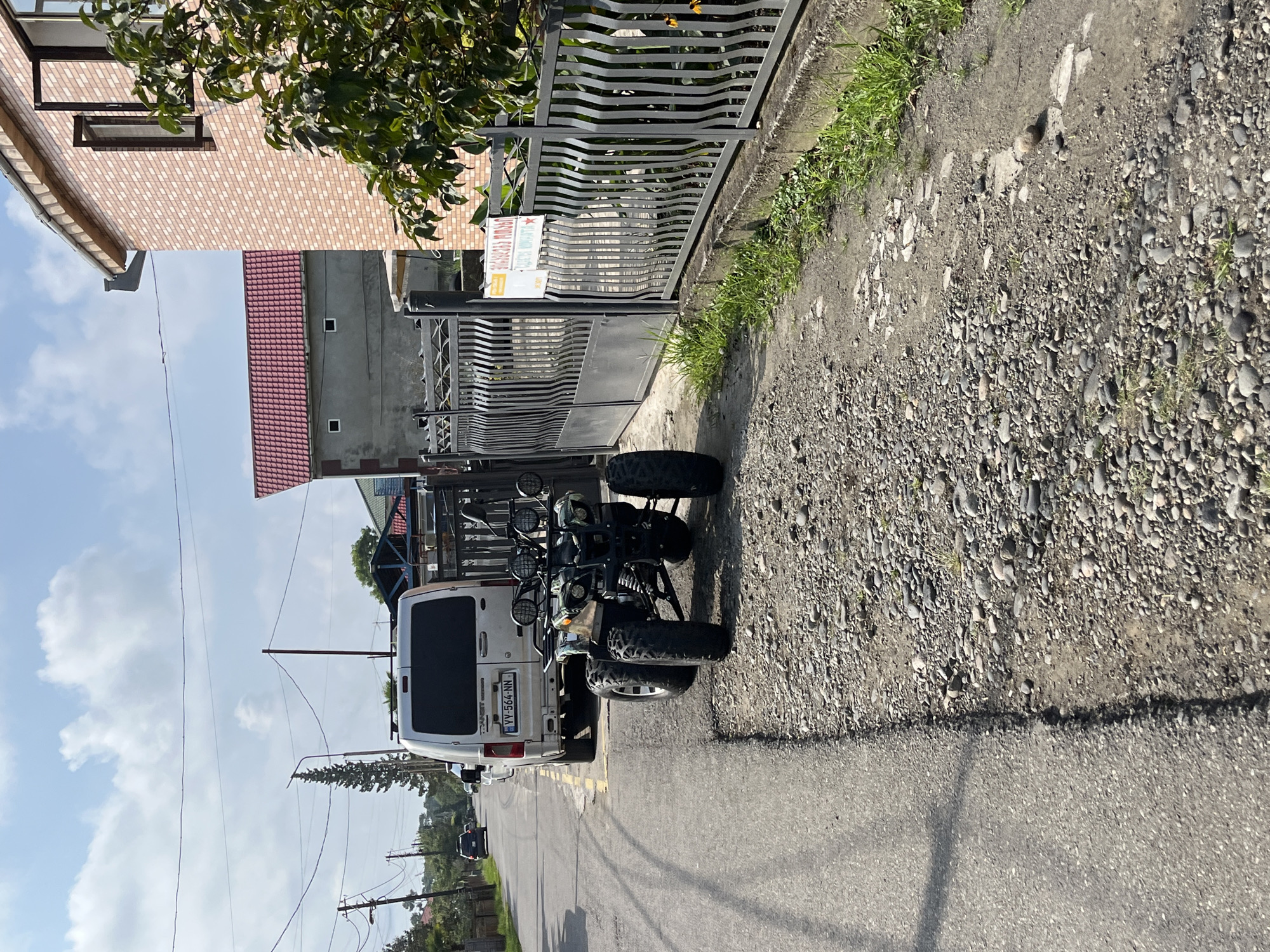 This is a street-level, eye-level, full-shot photograph taken in Ureki, Georgia, on a sunny day with scattered clouds. The scene depicts a narrow asphalt road with a gravel shoulder on the right. Parked on the shoulder, facing away from the camera, is a white van with the license plate "VY-564-NN." In front of the van and slightly to its right is a green and black all-terrain vehicle (ATV) with four large tires, also parked. Beyond the ATV and van, the road continues to recede into the distance, lined with houses and trees. To the right of the ATV and van is a metal fence, and behind it, a light pink brick building and a building with a red tiled roof are visible. A white sign with text in a non-English script is attached to the fence. The environment is rural or suburban, with evidence of both residential structures and a functioning roadway. There are no people visible in the image.