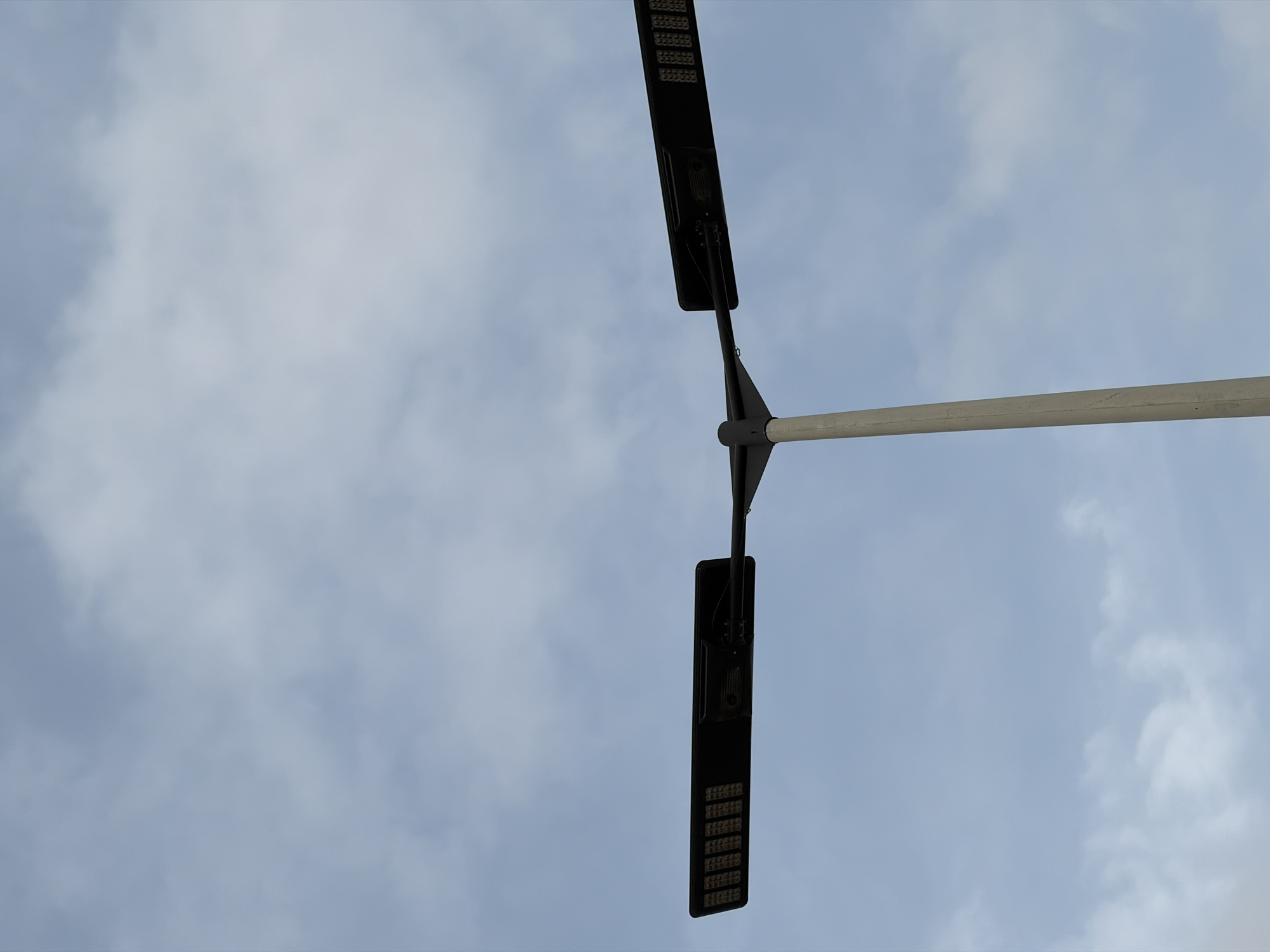 An upward-facing shot shows a grey concrete lamppost against a pale blue sky filled with wispy white clouds. The lamppost supports a horizontal fixture with two black, rectangular light heads on either end. Each light head contains multiple small, visible LEDs. The lamppost is located in Alaguntan, Nigeria.