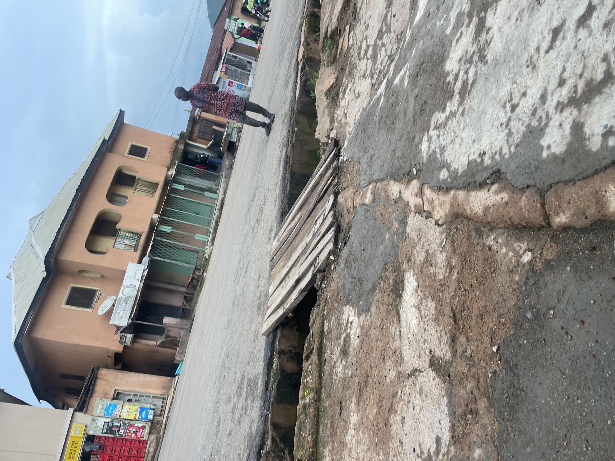 The image depicts a street scene in Jos, Nigeria. In the foreground, a cracked and weathered concrete surface transitions into a dirtier, more broken asphalt road with a gaping trench or gutter running alongside it. A makeshift wooden bridge, constructed from several planks, spans a portion of the trench.
In the mid-ground, a man wearing a traditional patterned dashiki and dark trousers stands on the asphalt road, facing away from the camera and looking towards the right. Behind him, several buildings line the street. One building, painted in a light terracotta color, has multiple stories, balconies, and awnings covering storefronts on the ground floor. These storefronts appear to be open with green metal grilles. A satellite dish is visible on the roof of this building. Signs, including one that reads "HAPPY FAST FOOD," are attached to the facade.
Further down the street, other buildings with corrugated metal roofs are visible, some with signage. A few other people can be seen near the storefronts, and a motorcycle is parked on the right side of the road. The sky above is overcast with a light blue and grey hue, indicating a cloudy day. The overall impression is of a typical urban environment with signs of wear and tear.