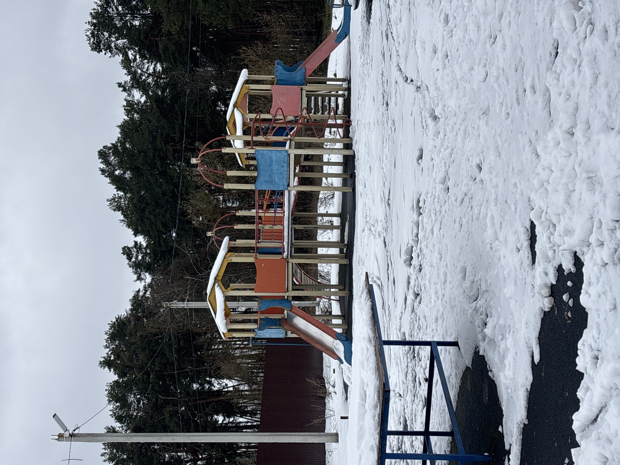 An outdoor scene captures a snow-covered children's playground during winter, likely in Раздолье, Russia. The photograph, taken during daytime under an overcast sky, features a multi-level play structure made primarily of wood with colorful elements in red, blue, and yellow. Slides, climbing bars, and platforms are visible, with the yellow roof sections partially covered in snow.
The ground surrounding the playground is heavily blanketed with thick, lumpy snow, showing some textured patterns from accumulation or previous disturbance. No people or ongoing activities are depicted, giving the scene a quiet, still atmosphere. In the background, a dense line of dark green evergreen trees suggests a forest or wooded area bordering the playground. A dark brown corrugated metal fence runs horizontally behind the lower part of the play structure, separating it from the treeline. To the left foreground, a utility pole with wires and what appears to be a security camera is visible. No text is discernible within the image.