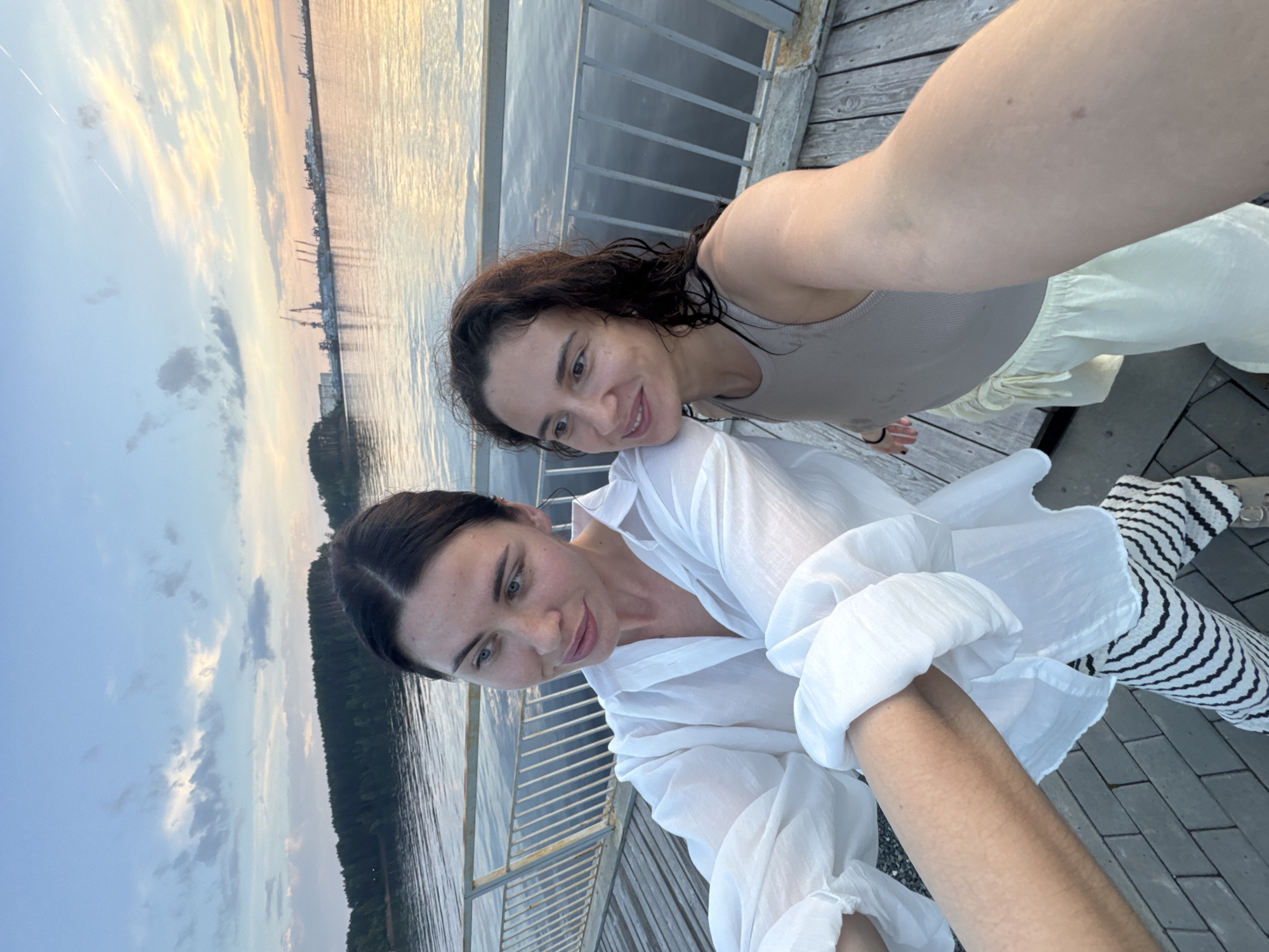Two women are taking a selfie on a wooden boardwalk near a body of water at dusk. The woman on the left is wearing a white oversized shirt and striped pants, and the woman on the right is wearing a light-colored tank top and light-colored shorts. The water is calm, reflecting the sky, which has soft clouds and a warm glow from the setting sun. In the distance, there is a treeline and a port with cranes and buildings. The boardwalk has a metal railing. This location is likely along the Kama River in Chaikovsky, Russia, given the presence of a port and the overall landscape.