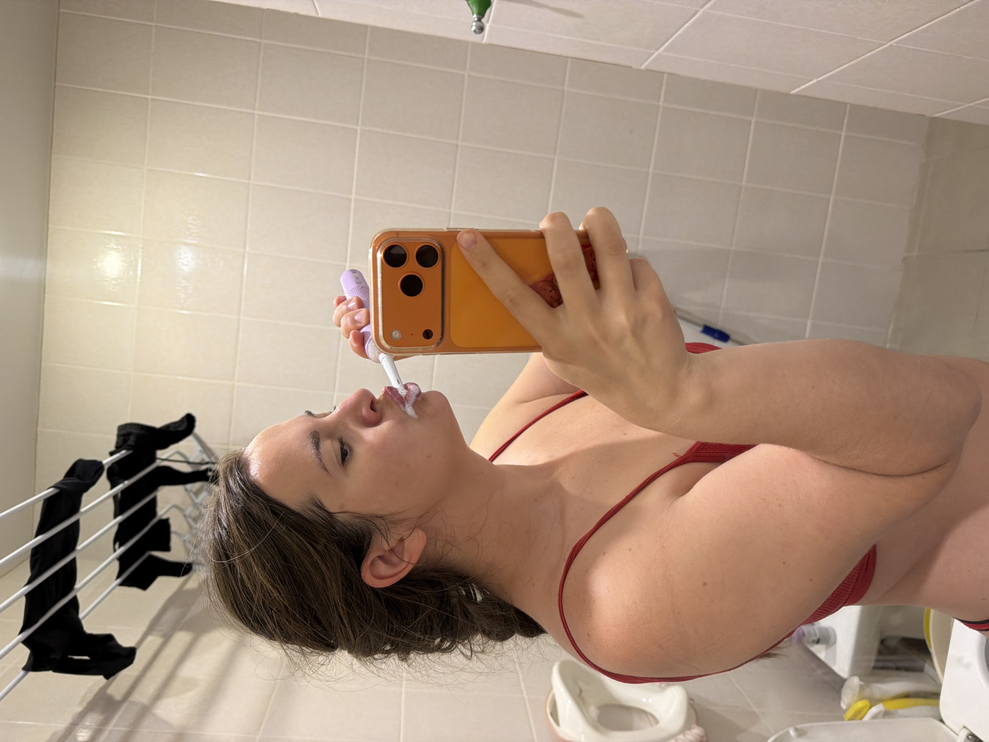 A woman in a red top is brushing her teeth while taking a selfie in a bathroom. The bathroom has beige tiles, a clothes drying rack with black clothes, and various bathroom fixtures and items visible in the background. There is no information to determine that this image was taken in the city of Sochi, Russia.