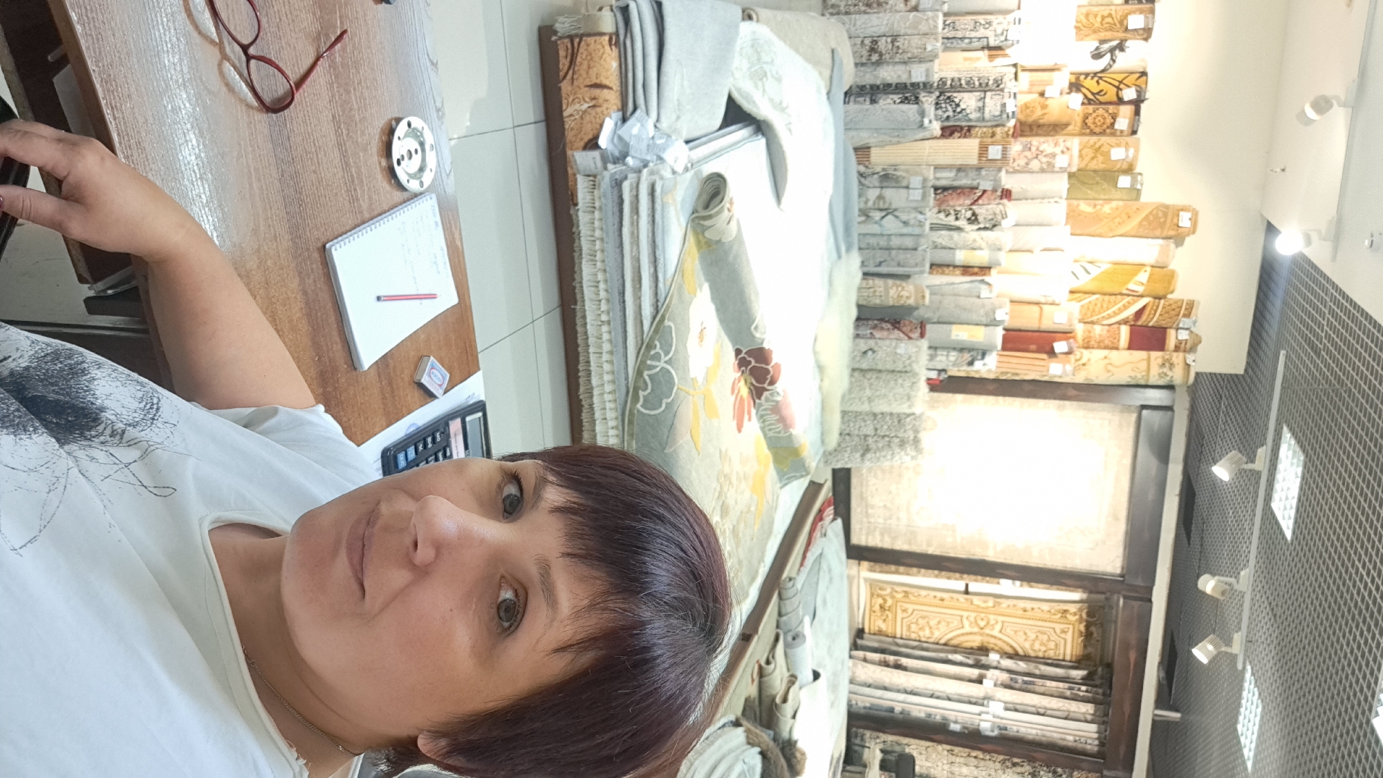 A woman with short, dark hair and brown eyes is taking a selfie in what appears to be a carpet store. She is wearing a white t-shirt. On the wooden desk in front of her are a calculator, a notepad with a red pencil, a matchbox, and a pair of red-rimmed glasses. In the background, stacks of rolled-up carpets are displayed on shelves. The store has a tiled floor and a suspended grid ceiling with track lighting. The location is identified as Barnaul, Russia.