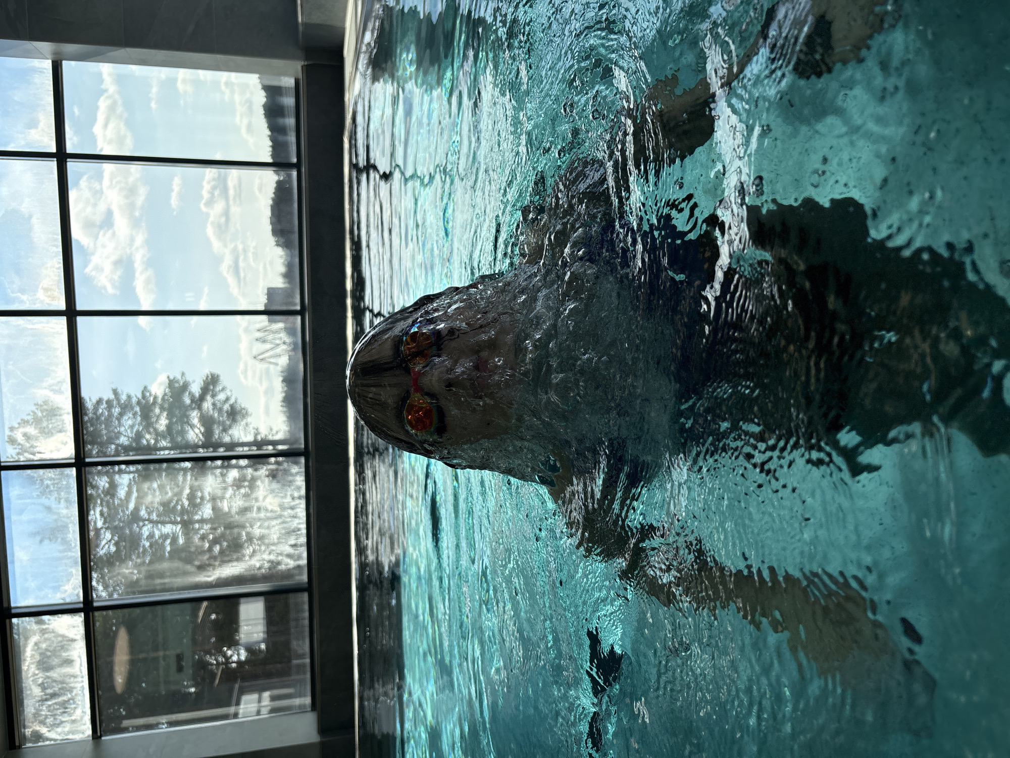 A person is swimming in an indoor pool. The person is wearing goggles and their head is above the water. Bubbles and ripples are visible in the water around the person's head. A large window is visible on the left side of the frame, showing a view of trees and a cloudy sky. The pool appears to be located inside a building, possibly in Chaikovsky, Russia.
