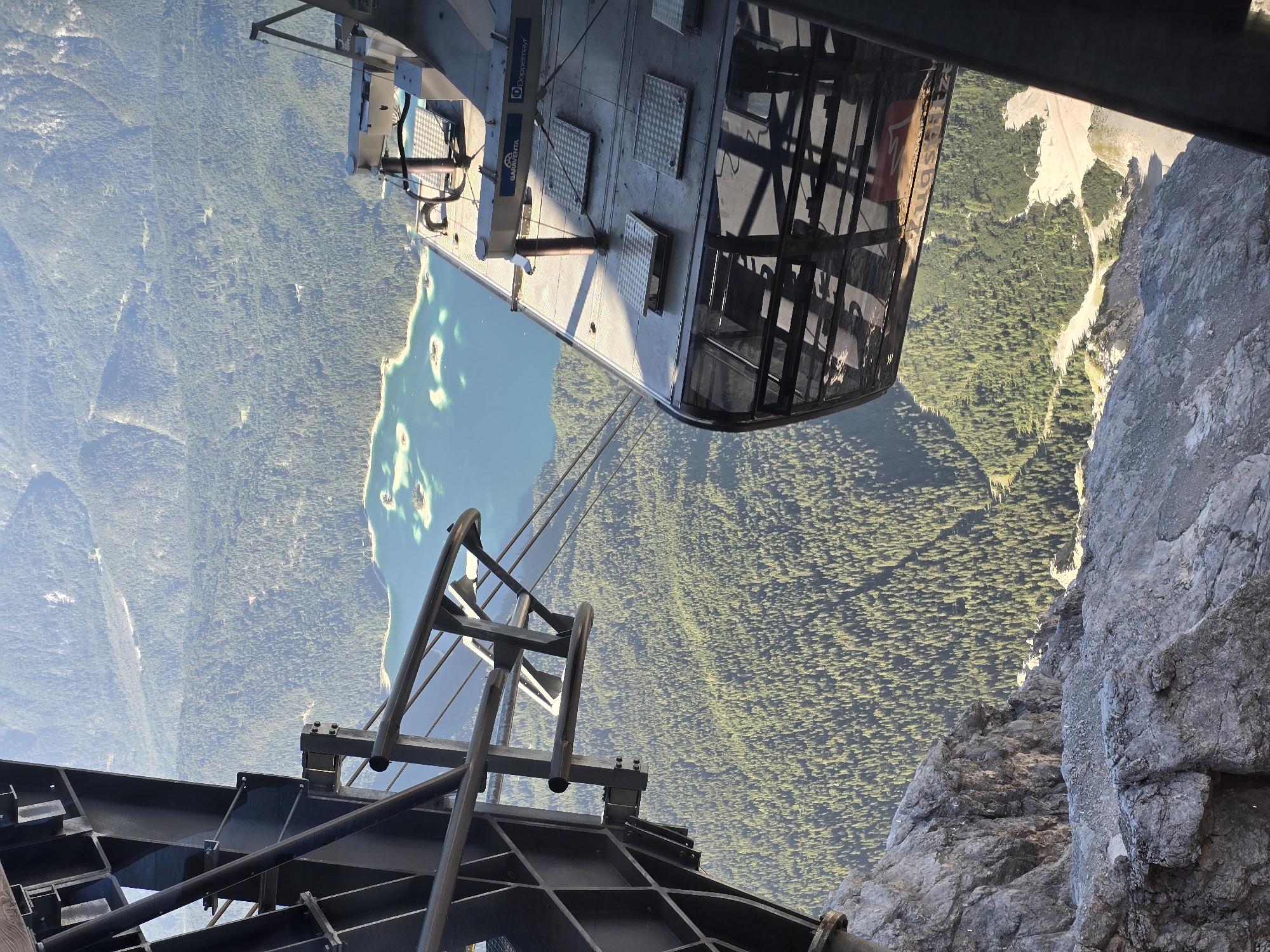 This image captures a view from a cable car ascending a mountain, likely the Zugspitze, near Garmisch-Partenkirchen, Germany. The cable car, a modern design with large windows and metallic components, is positioned in the foreground, partially obscuring the dramatic drop below. The view through the cable car windows and over its roof reveals a vast expanse of lush green forest blanketing rolling mountain slopes. In the mid-ground, a striking turquoise lake with several small islands is visible, its calm surface reflecting the sunlight. The sky is clear and bright, suggesting a sunny day with excellent visibility. The rocky, mountainous terrain at the bottom of the frame hints at the high altitude and rugged landscape. The overall scene conveys a sense of awe-inspiring natural beauty and the thrill of mountain exploration.