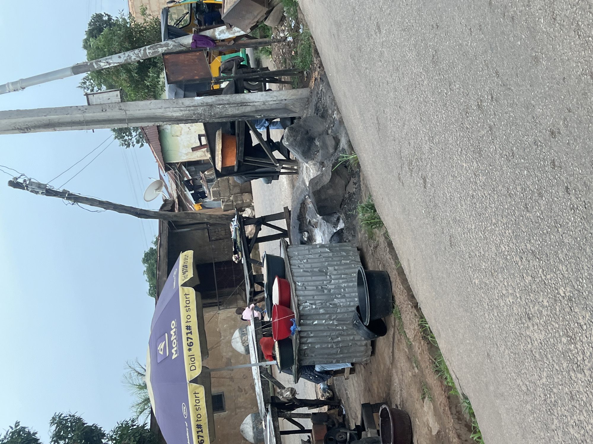 The image depicts an outdoor market stall in Jos, Nigeria, with a focus on the sale of grains, likely rice, which are contained in large bowls. A purple umbrella with "MoMo" branding and "Dial *671# to start" is set up, providing shade over the stall. Several large black and red bowls are visible, some filled with white grain. A corrugated metal structure is used as the base for the display. Behind the stall, a child in a pink shirt is seated. Various other market elements are present, including wooden tables, makeshift structures, a motorcycle engine, and a tricycle. The ground is paved with asphalt, and the scene is set against a backdrop of simple buildings and electrical poles, suggesting a typical urban or semi-urban environment in Jos. The sky is overcast, indicating possibly cloudy weather.