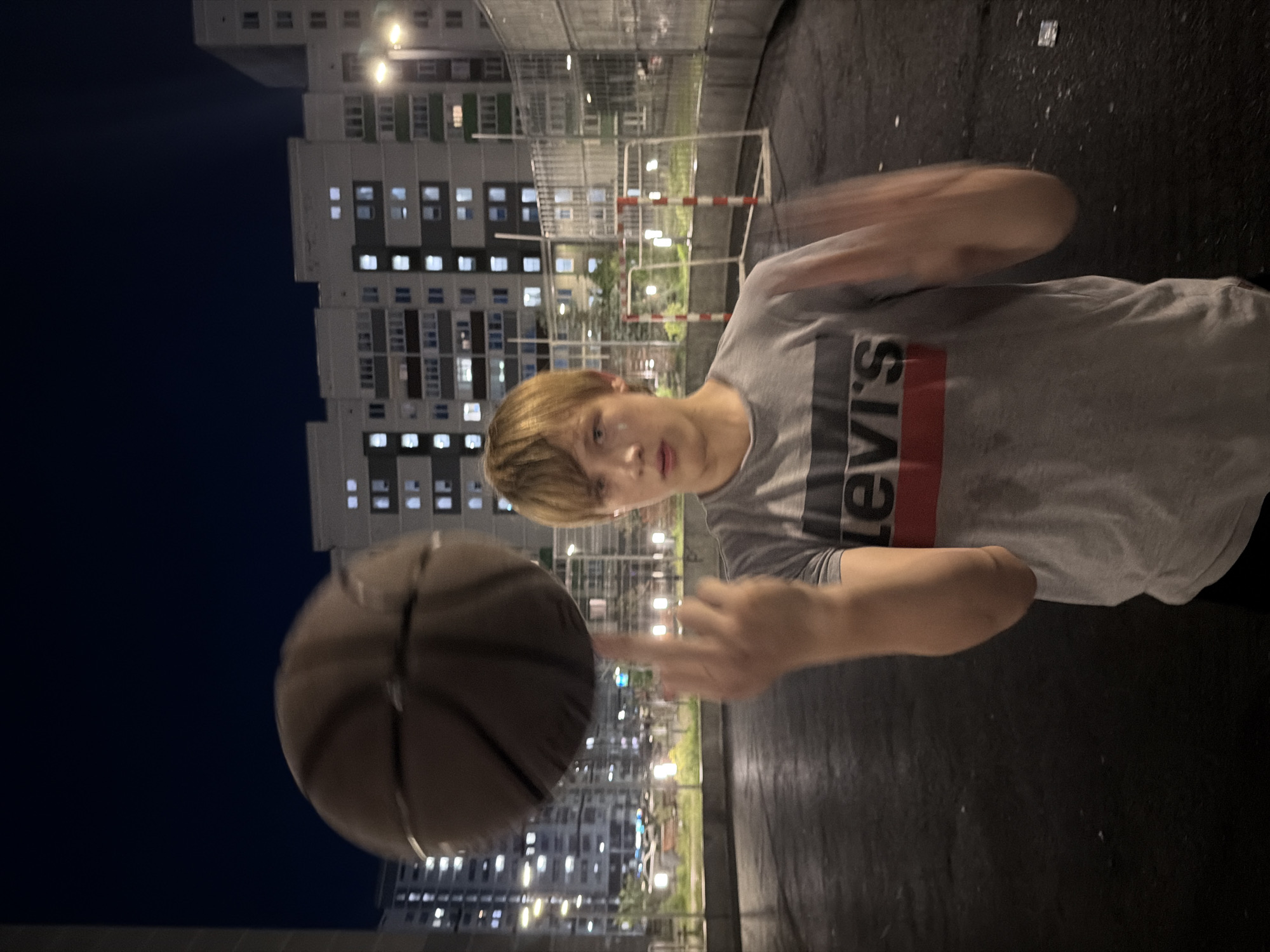 A young Caucasian male with light brown hair is outdoors at night, balancing a basketball on his right index finger. He is wearing a grey t-shirt with a red and black horizontal stripe design and the word "LEVI'S" printed in white on the front. The background shows apartment buildings with illuminated windows and streetlights, suggesting an urban setting in Izhevsk, Russia. The ground appears to be a dark, paved surface, possibly a basketball court. In the mid-ground, there is a soccer goal with red and white posts. The overall lighting is dim, typical of nighttime in a city.
