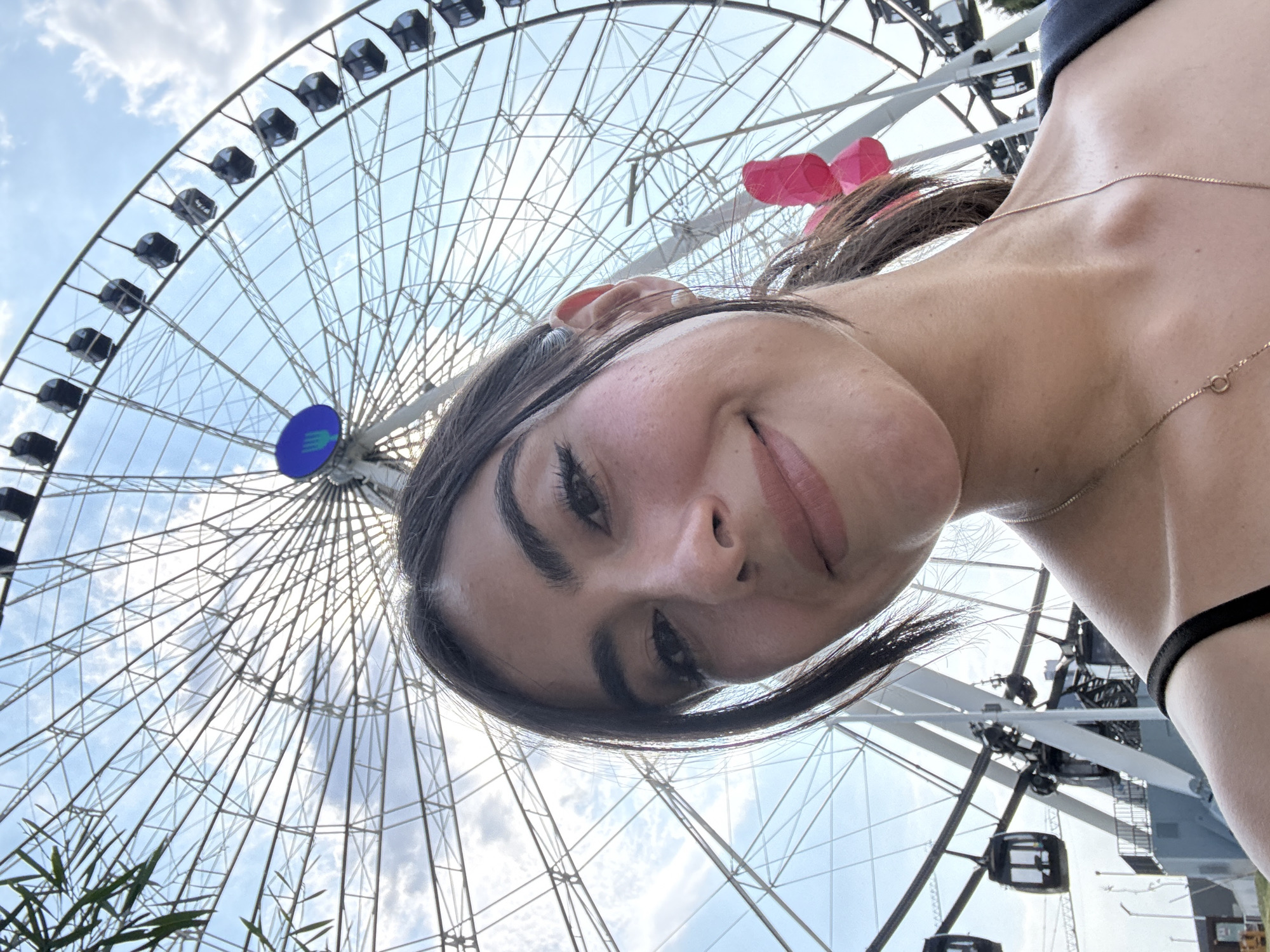 A young woman is taking a selfie with a Ferris wheel in the background. The Ferris wheel is large and white with black gondolas. The sky is blue with some clouds. The woman has dark hair and eyebrows, light skin and is wearing a thin gold necklace. She is smiling and looking at the camera. The photograph appears to be taken from a low angle. The photograph was most likely taken in Puebla, Mexico.