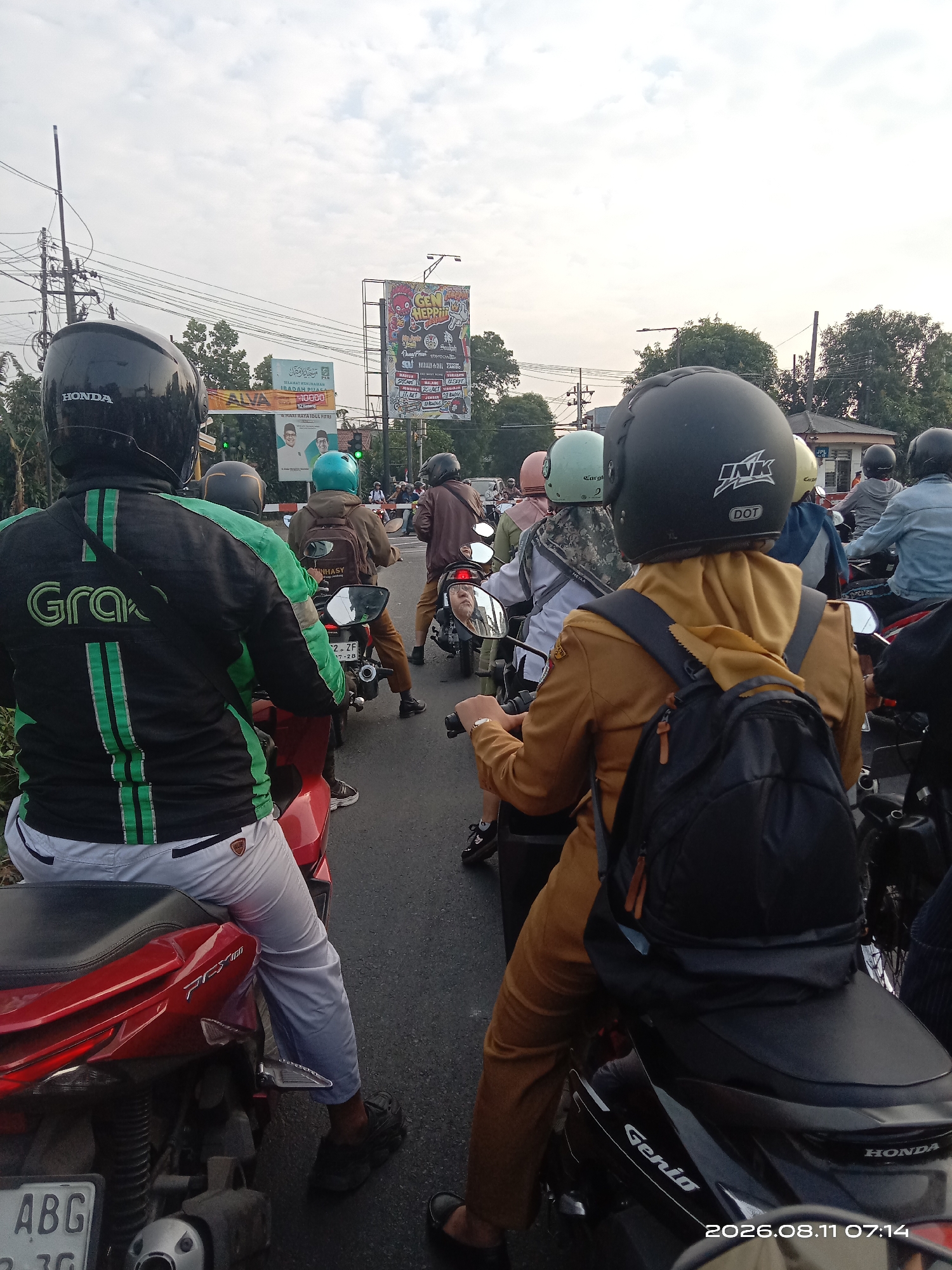 The image captures a traffic jam scene in Jabon, Indonesia, likely during the late afternoon given the lighting. Multiple motorcycles are queued on a paved road, suggesting a busy urban or suburban thoroughfare. The sky is overcast with a light gray hue. On the right side of the road, there is a building with a light-colored facade and a dark roof, behind which trees are visible.
In the foreground, a person wearing a black helmet with "HONDA" branding is seated on a red motorcycle, facing away from the camera. They are wearing a black and green jacket with "Grab" logos on the back and sleeves, and light gray pants. To their right, another person is seated on a motorcycle, also facing away, wearing a tan uniform with a black backpack. Their helmet is black with "INK" and "DOT" labels.
Further in the middle ground, several other motorcyclists are visible, some wearing helmets of various colors, including green and blue. One individual in the middle appears to be wearing a brown jacket and brown pants. Beyond the motorcycles, several billboards are erected along the roadside. One prominent billboard features colorful graphics and text, another displays an image of two men in suits with text in Indonesian, and a third appears to be a banner with Indonesian text. Utility poles with numerous wires are also present along the roadside, indicating infrastructure typical of Indonesian towns and cities.
The overall impression is one of congestion and daily life in a bustling Indonesian locale. The presence of Grab branding suggests this is a common mode of transportation and delivery service in the area.