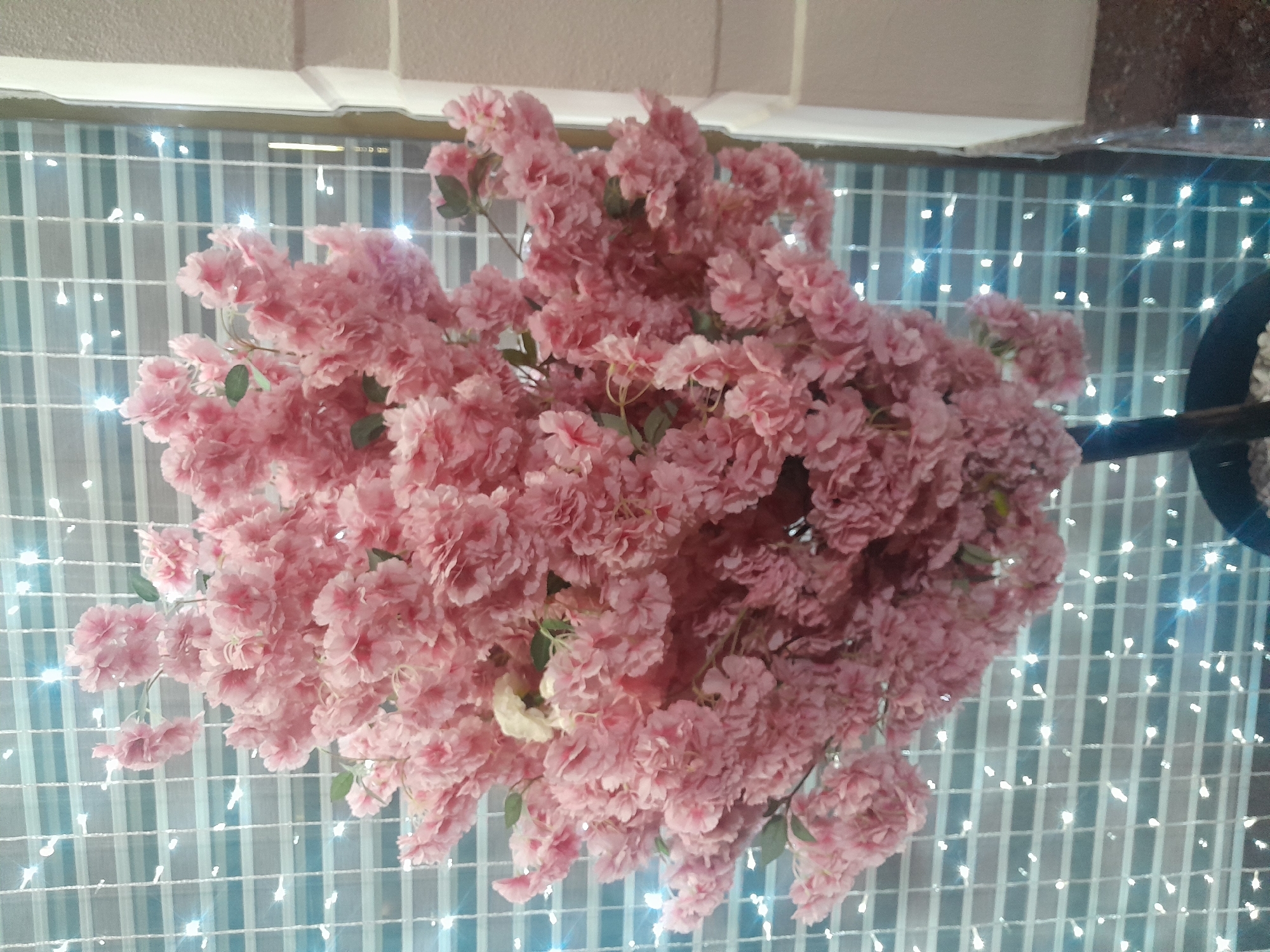 An artificial cherry blossom tree with pink flowers is positioned in front of a window with horizontal white blinds and small, twinkling lights. The tree has a dark brown trunk and is planted in a black pot filled with white pebbles. The background consists of a grid pattern formed by the blinds and the scattered lights, which are bright white and appear to be embedded in or behind the blinds. To the right of the tree, a section of a cream-colored wall with a textured surface is visible, along with a darker marbled countertop or ledge. The lighting in the scene is soft, and the focus is primarily on the dense cluster of pink blossoms.