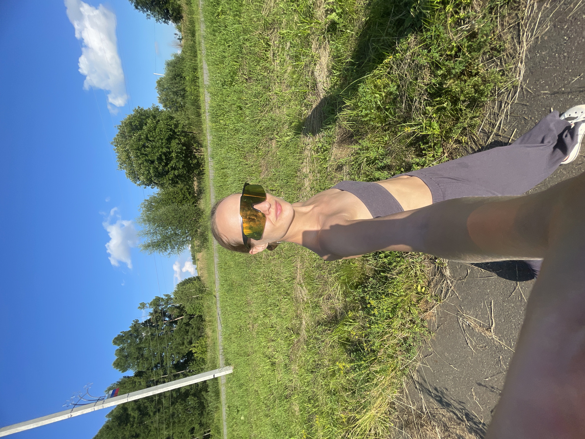 A woman wearing a grey crop top and grey wide-leg pants, along with oversized sunglasses, stands outdoors. She is holding her phone in a selfie pose. The background consists of a grassy field and trees under a blue sky with clouds. A utility pole with a Russian flag attached is visible to the left. The scene appears to be on the outskirts of Podshivalovo, Russia, with a paved surface in the foreground.