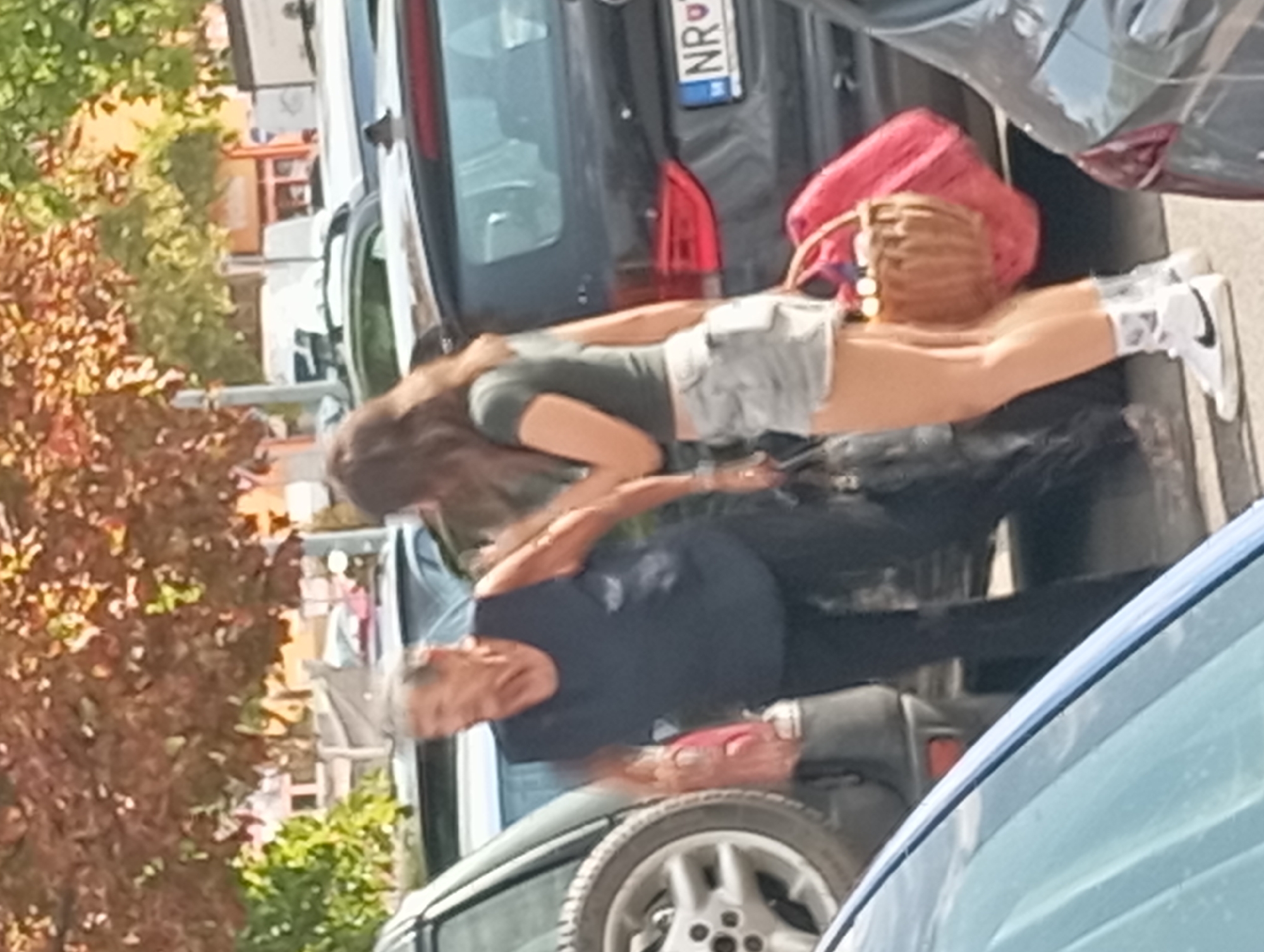 Two women are standing next to parked cars in what appears to be a parking lot. The woman on the left, an elderly individual with gray hair, is wearing a dark sleeveless shirt and black pants. She is holding a red bag in her right hand and is looking to her right. The woman on the right, who is younger, has long brown hair and is wearing a green t-shirt and light-colored shorts. She is carrying a wicker basket in her left hand, with a large red bag slung over her left shoulder. She is looking towards the left. The background consists of trees with autumn foliage and buildings. The license plate of the car to the right of the younger woman appears to be from Nitra, Slovakia, indicated by "NR" on the plate.
