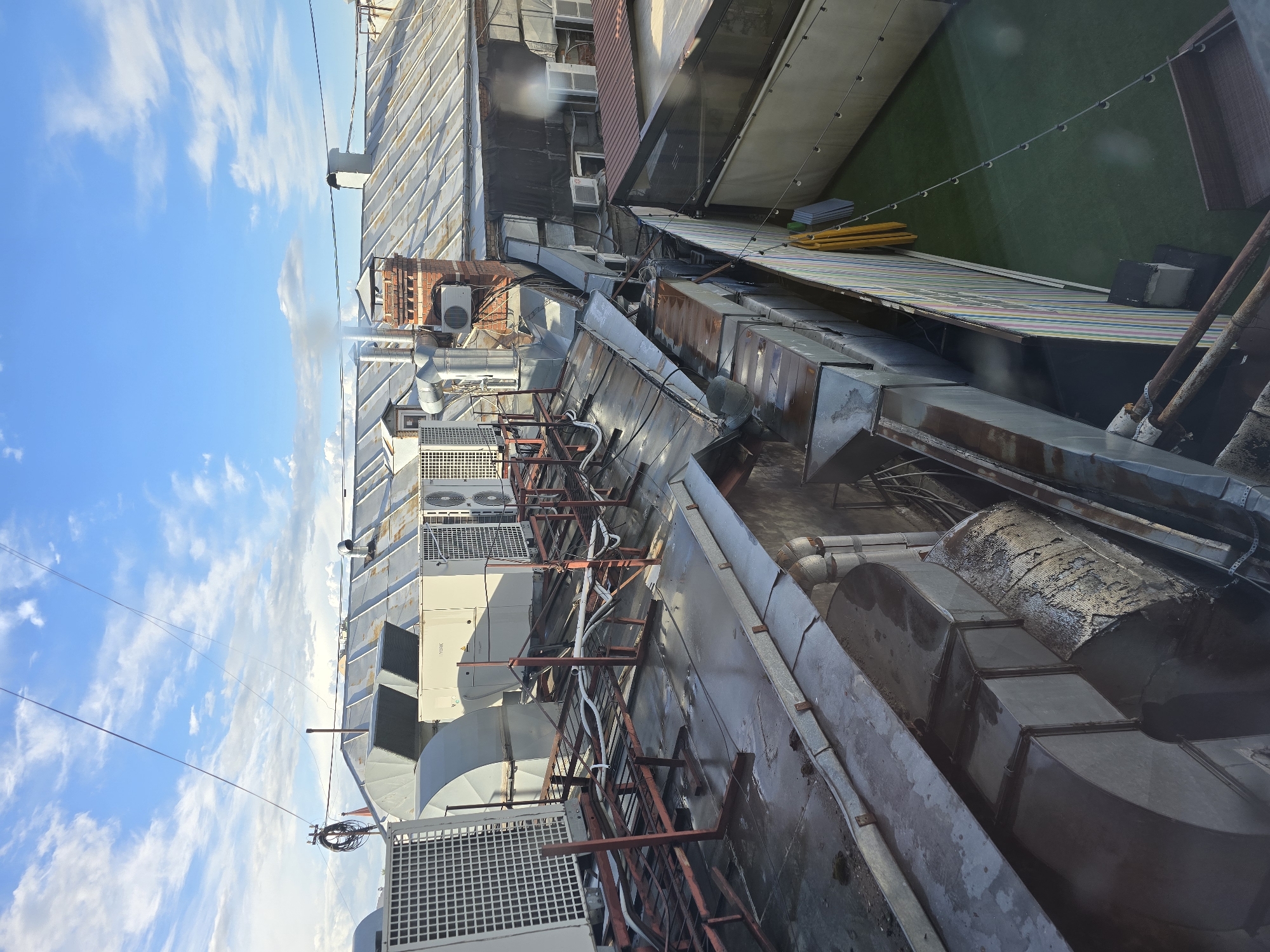 The image shows a rooftop in Moscow, Russia, densely packed with industrial infrastructure. Multiple air conditioning units are visible, interspersed with ventilation ducts and metal conduits. The metal surfaces show signs of age and weathering. The background features other buildings, including one with a green artificial turf surface. The sky is partly cloudy.